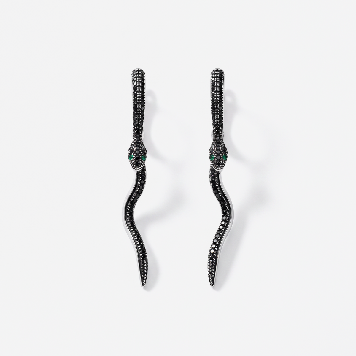 A pair of snake-shaped drop earrings in black metal with small green gemstone eyes, shown on a clean light gray background.