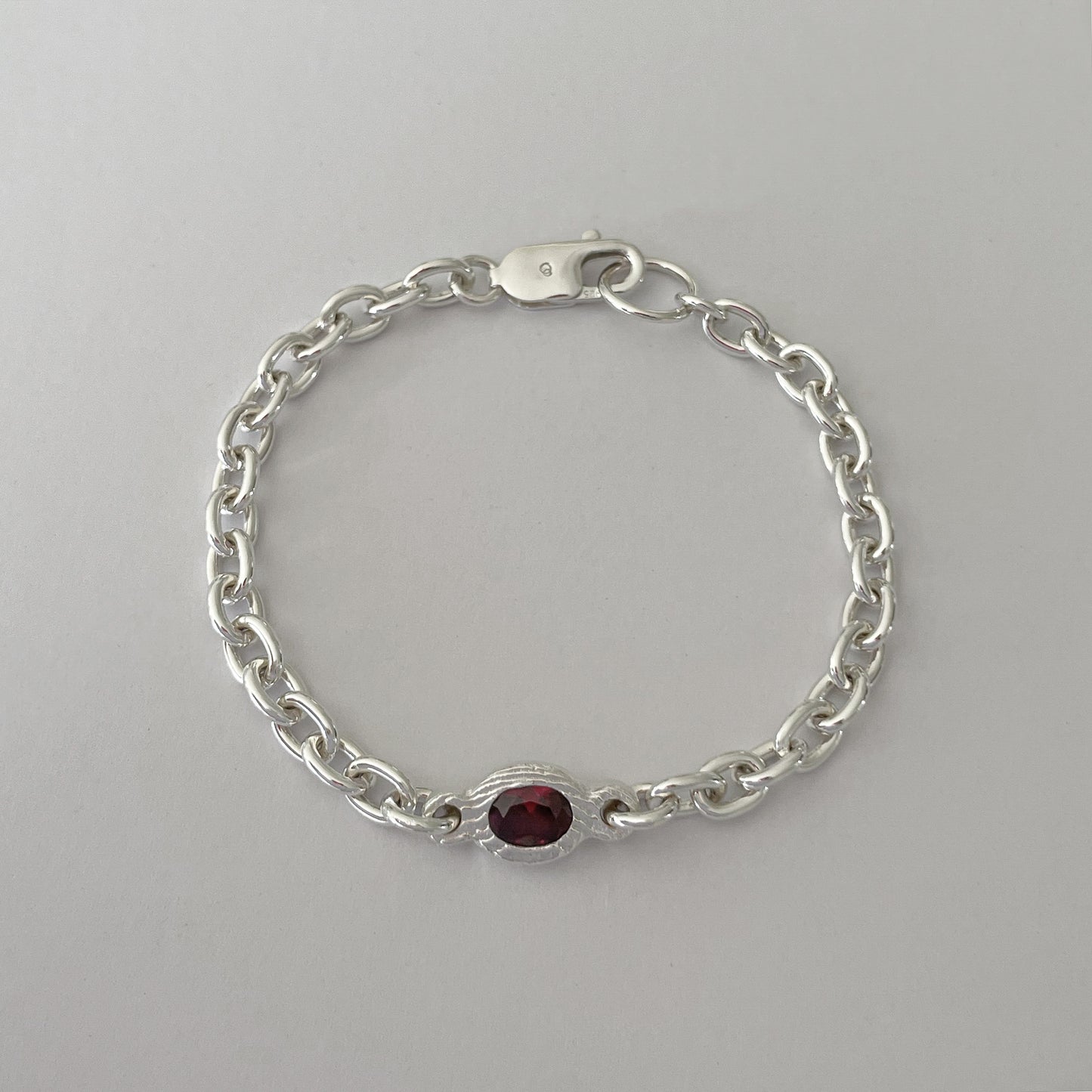A silver chain bracelet with a small oval red gemstone centerpiece, displayed on a neutral light gray background.