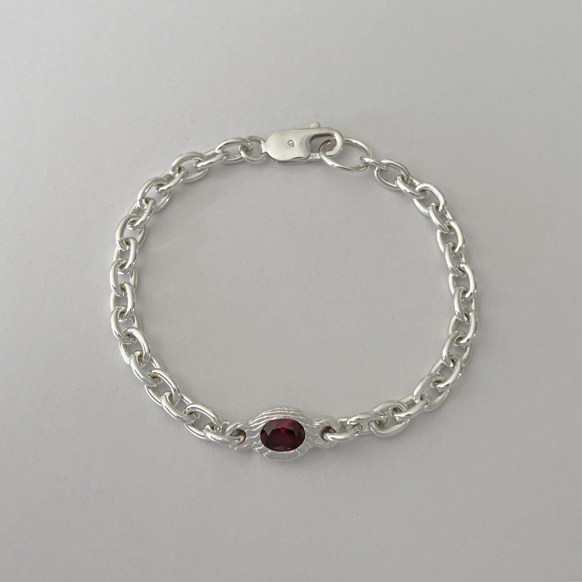 A silver chain bracelet with a small oval red gemstone centerpiece, displayed on a neutral light gray background.