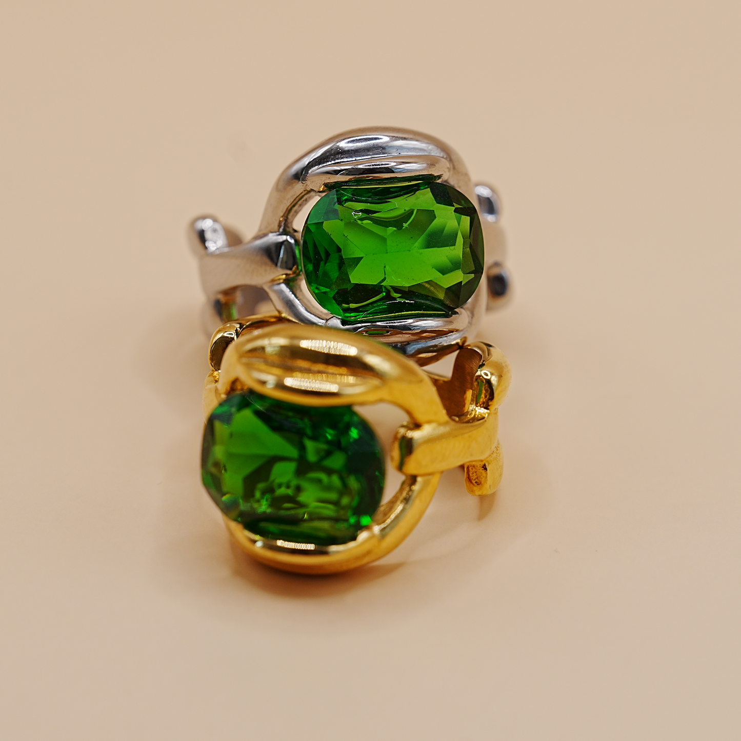 Two rings with large green faceted gemstones in silver and gold settings, photographed on a plain beige background.