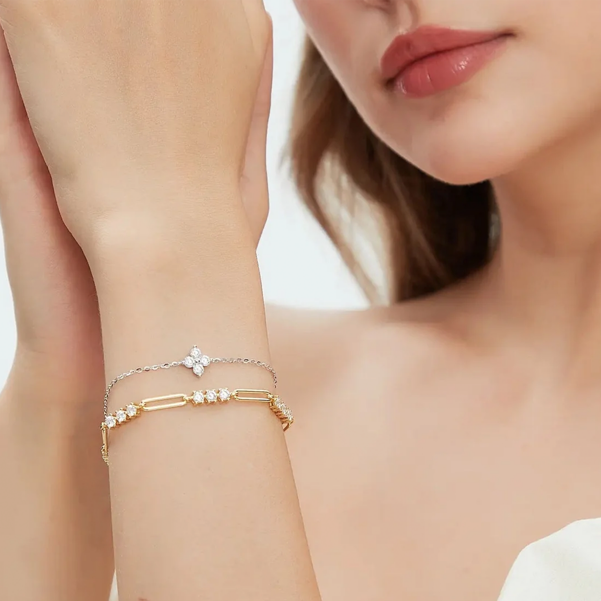 A close-up lifestyle shot of a model's wrist wearing two delicate bracelets: a silver chain bracelet with a small white flower charm and a gold bar-link bracelet adorned with small clear stones.
