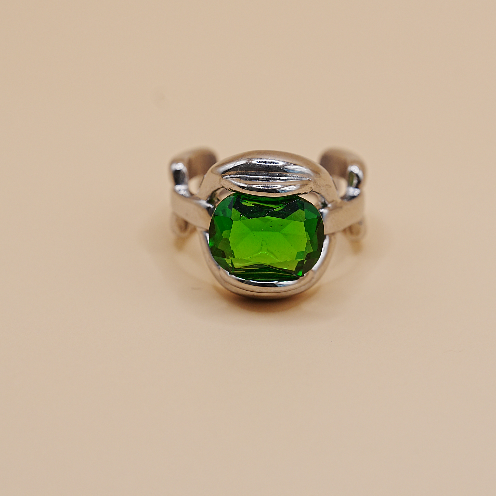 A silver ring featuring a large, cushion-cut green gemstone in a polished bezel setting, shown against a solid beige background.