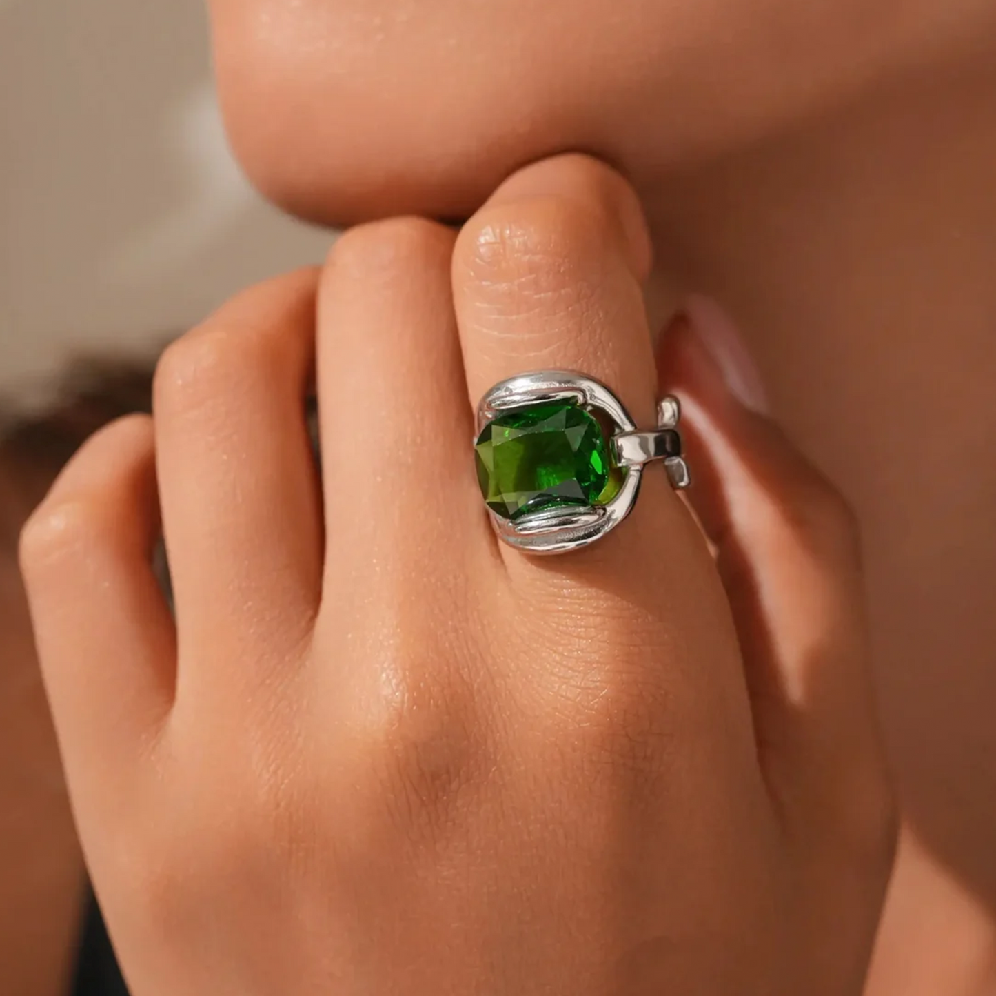 A close-up of a hand wearing a silver ring with a faceted green gemstone, set against a softly blurred skin-toned background.
