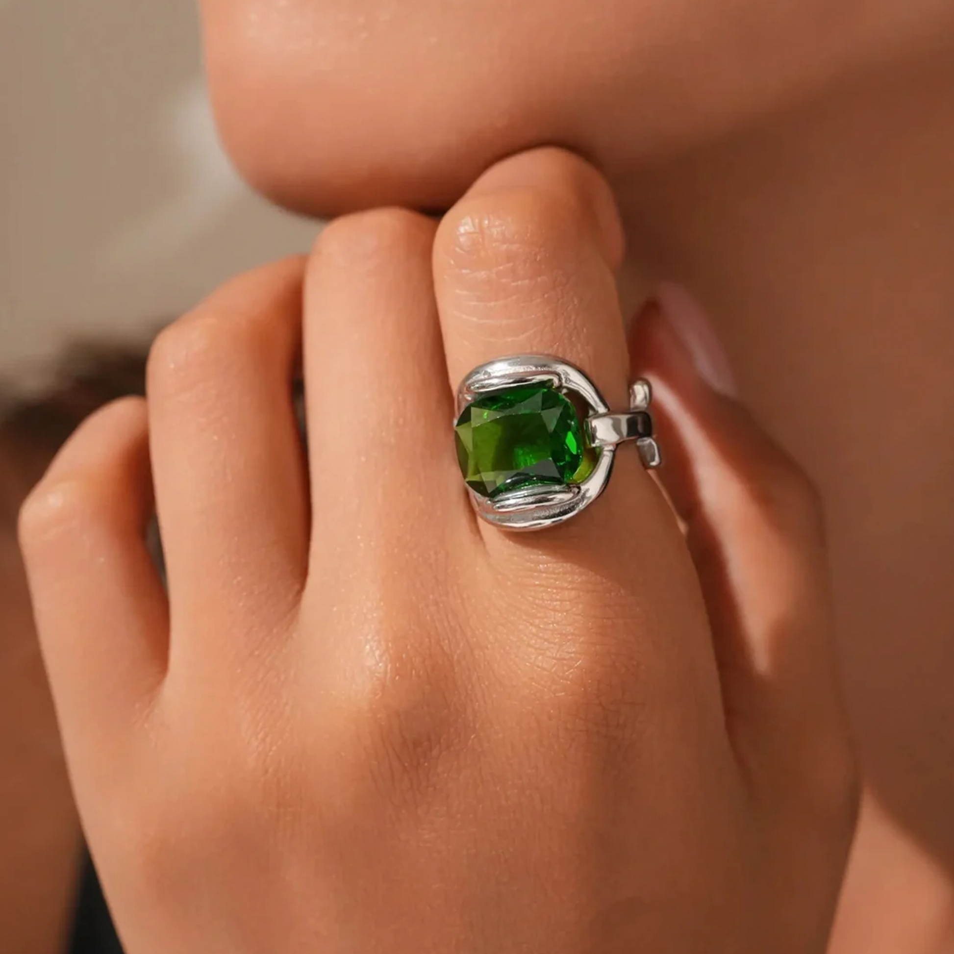 A close-up of a hand wearing a silver ring with a faceted green gemstone, set against a softly blurred skin-toned background.