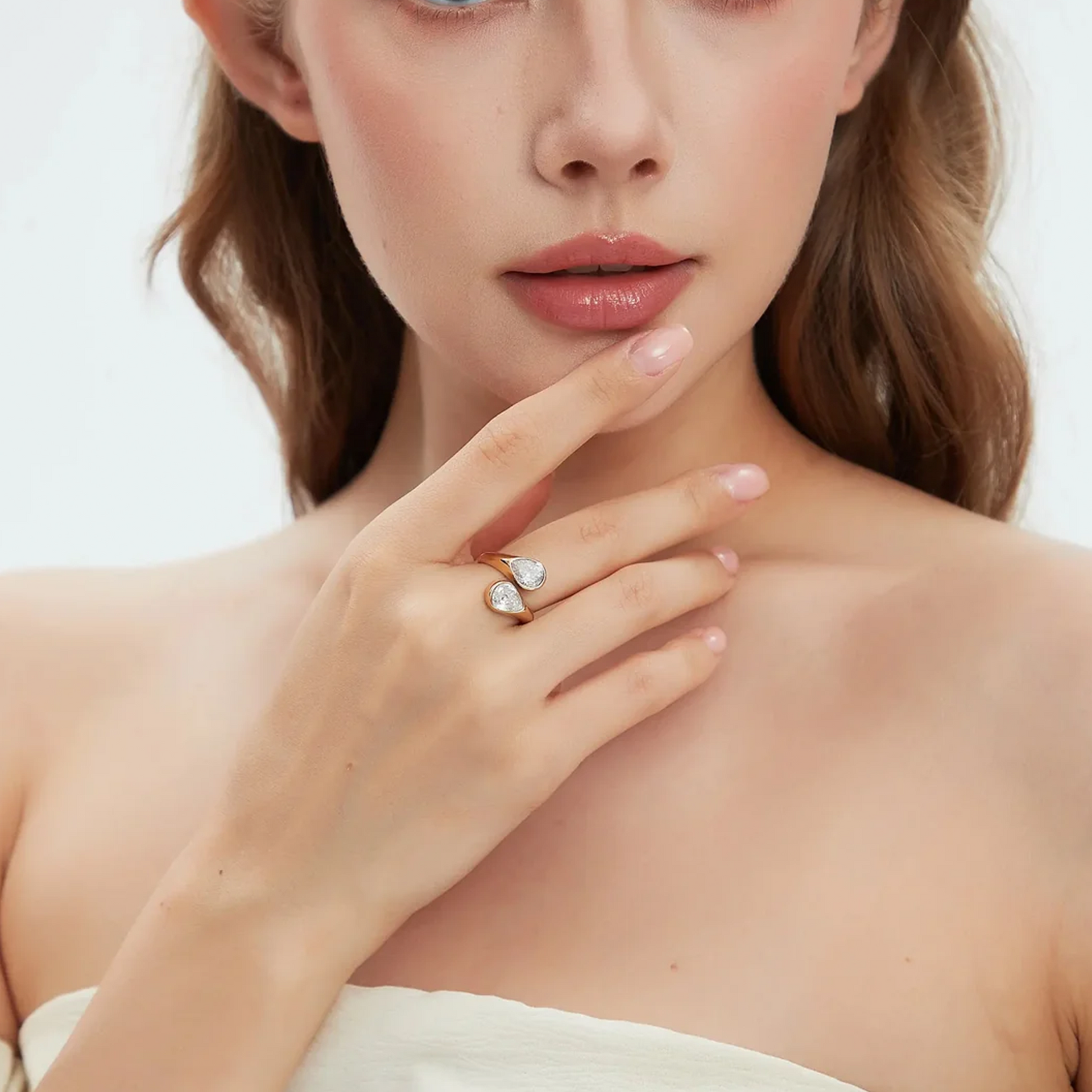 A close-up of a woman wearing a gold ring with two teardrop-shaped clear gemstones, resting her hand near her chin against a soft light background.