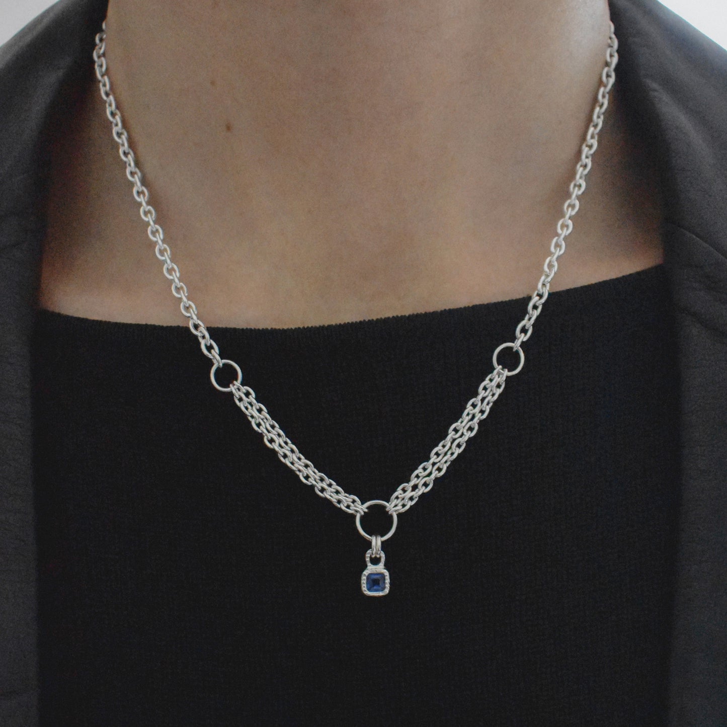 A silver chain necklace with a small blue gemstone pendant, shown worn on a person's neck over a black top.