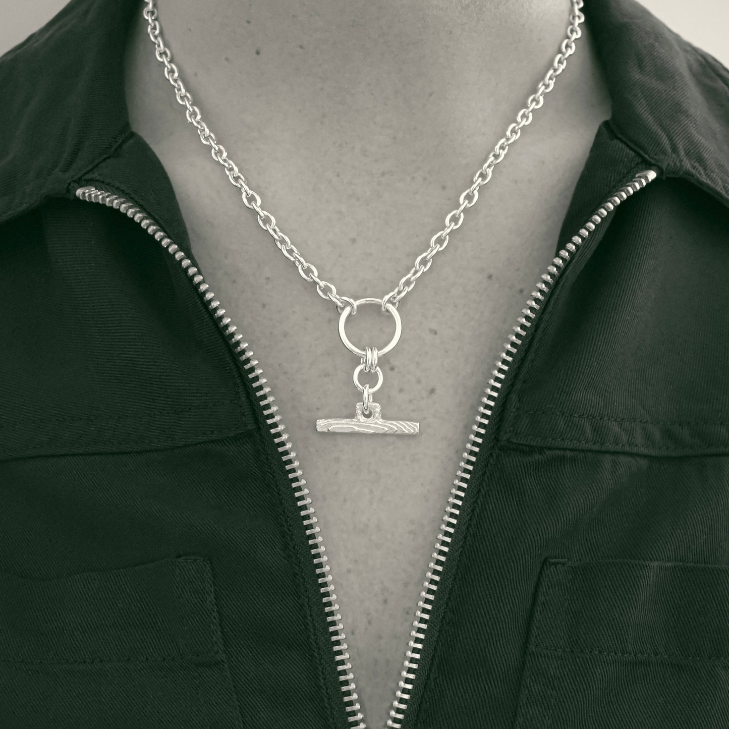 A silver-toned chain necklace with a circular ring and a small horizontal bar pendant, displayed on a mannequin torso wearing a dark zip-front jacket.