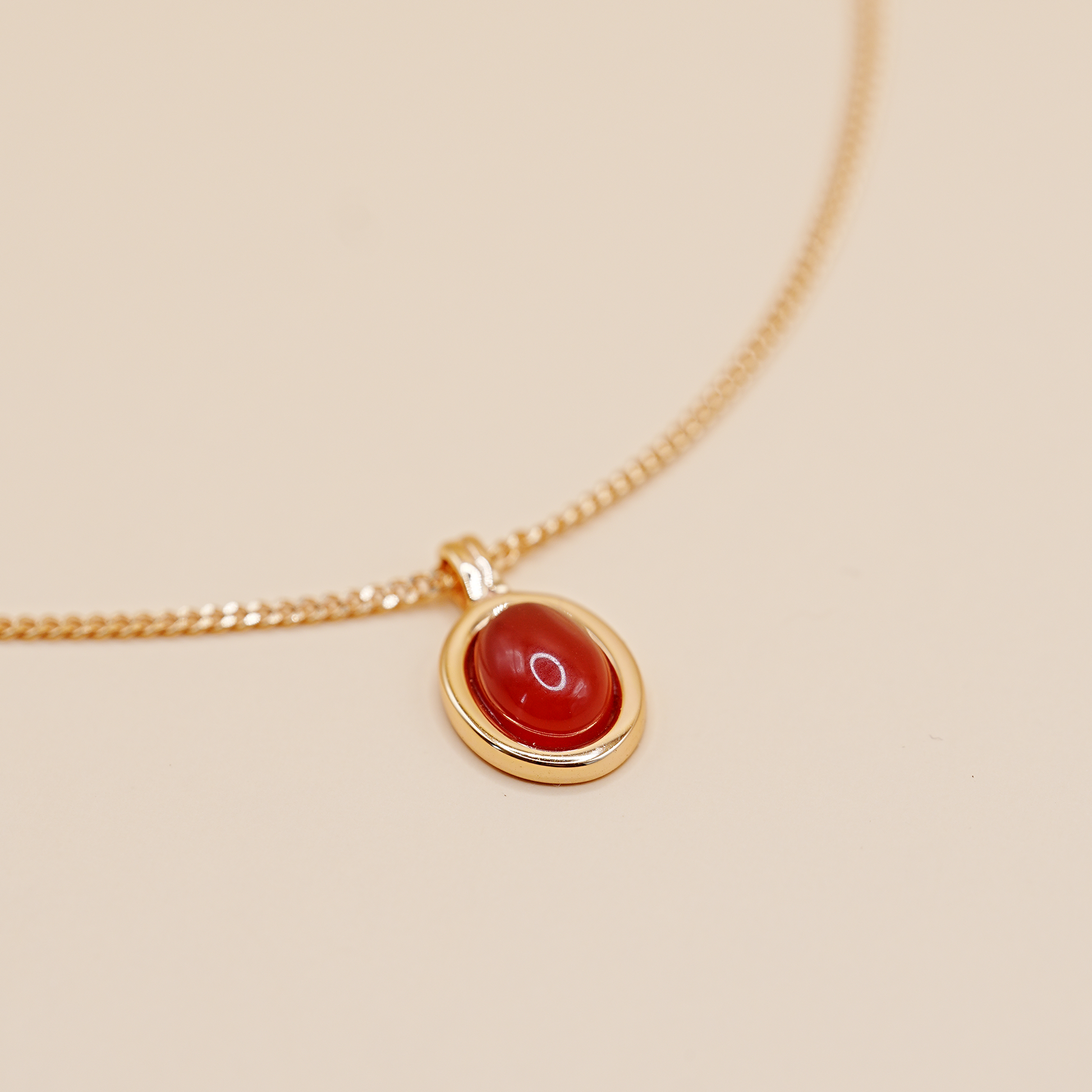 A gold chain necklace with an oval red cabochon gemstone pendant, displayed on a warm beige background.