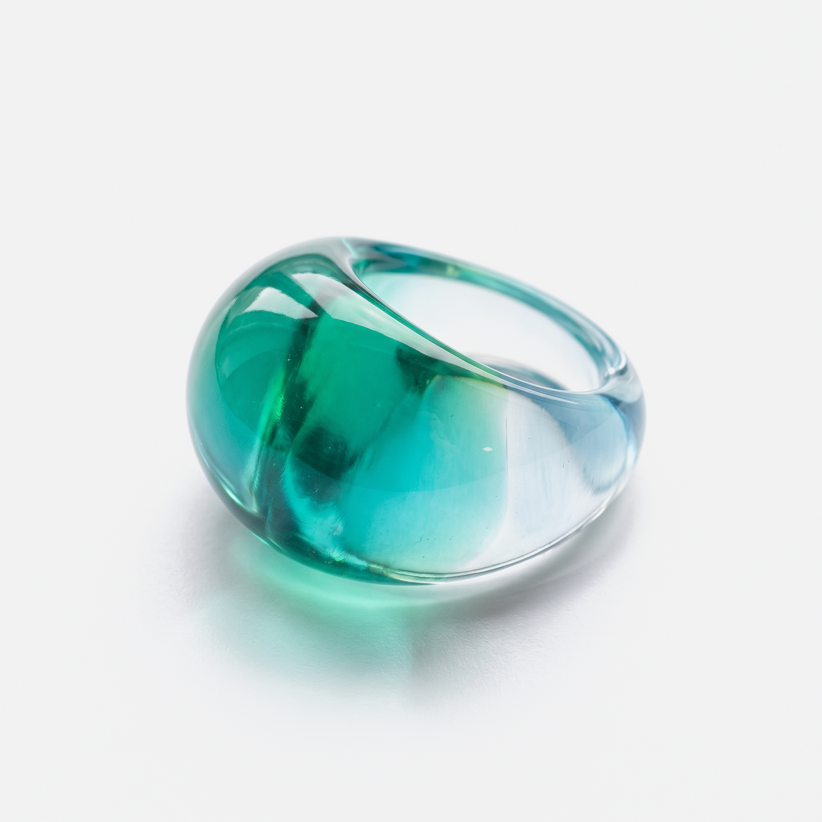 A translucent glass ring with a rounded, organic shape, featuring a gradient of teal green and light blue, photographed on a clean white background.
