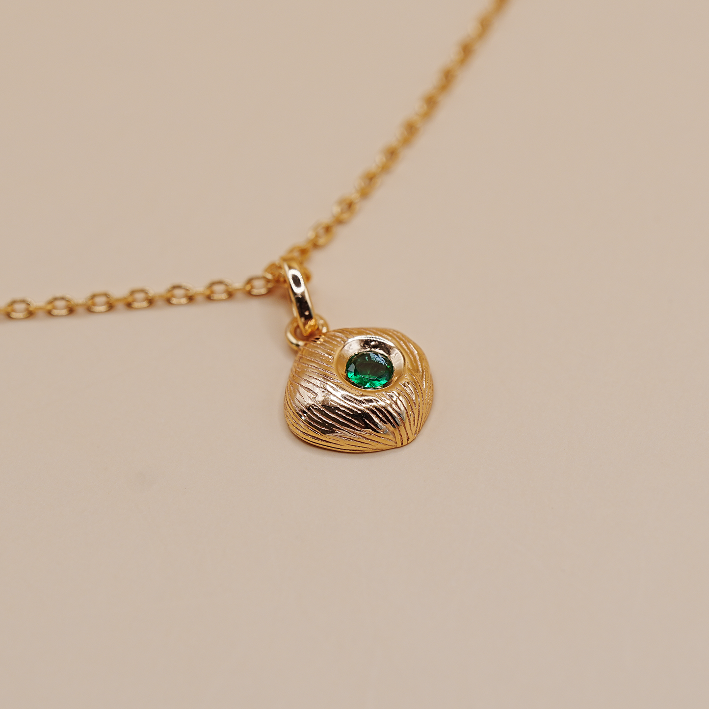 A gold pendant necklace featuring a textured, organic-shaped pendant with a small, round green gemstone inset, shown on a delicate gold chain against a solid beige background.