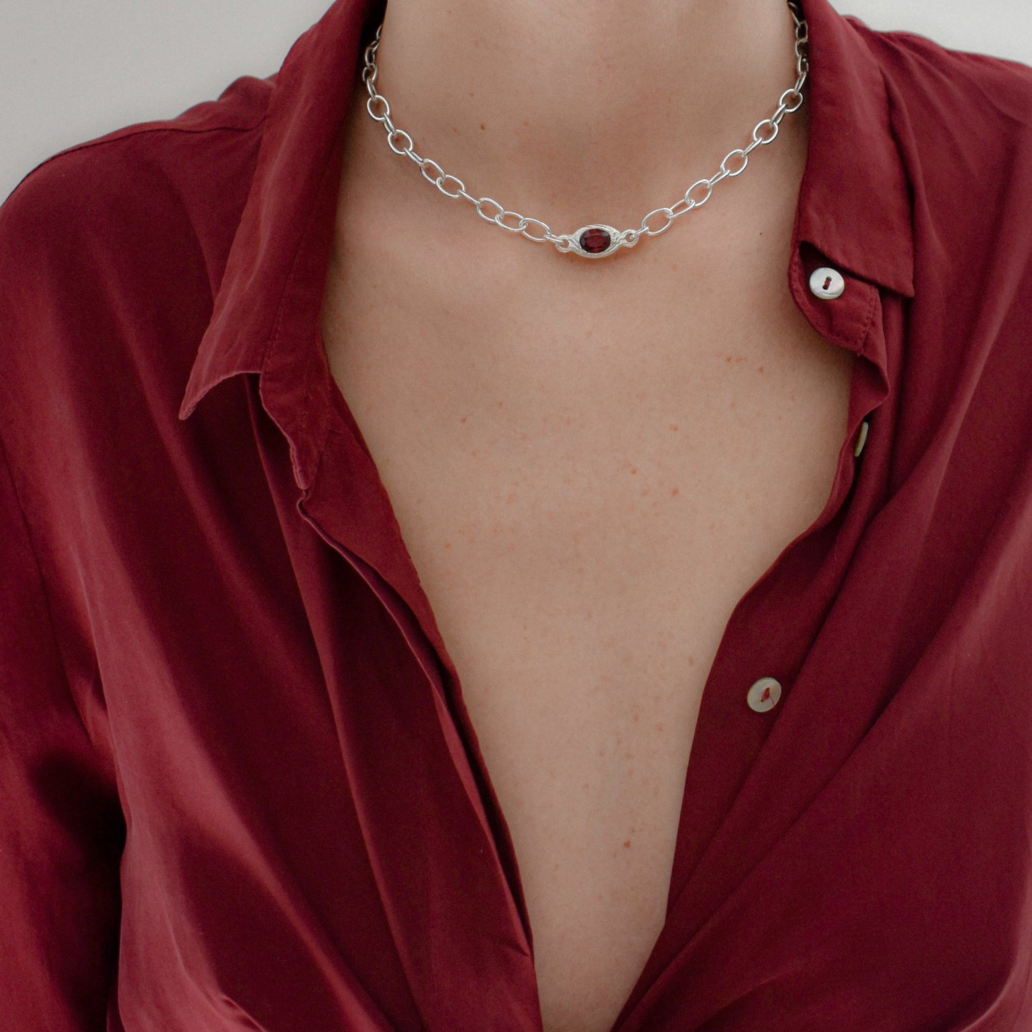 A close-up of a silver chain necklace with a central oval dark red gemstone, worn by a model wearing a deep red satin button-down shirt.