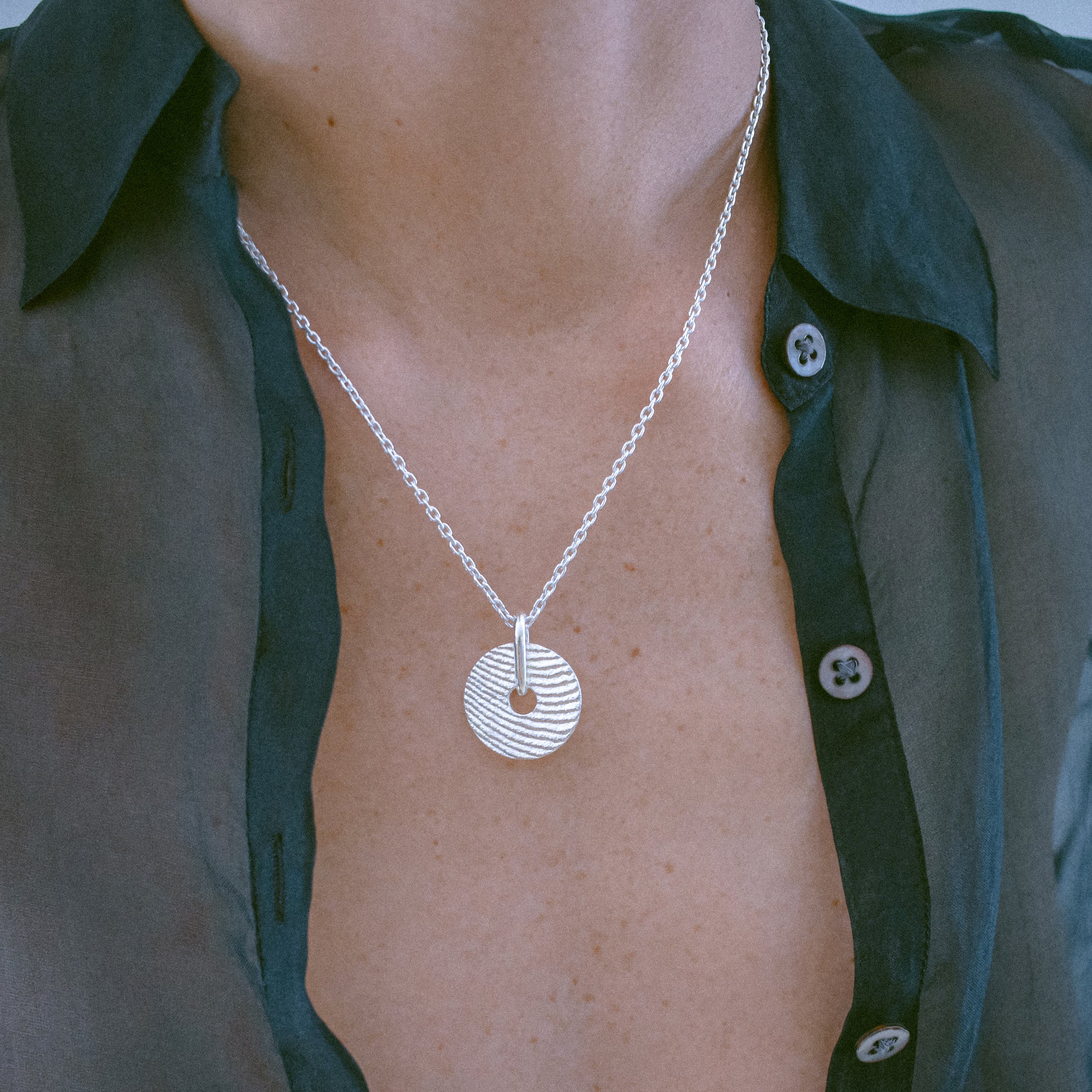 A silver pendant necklace with a textured circular disc design, worn by a person wearing a dark green button-down shirt.