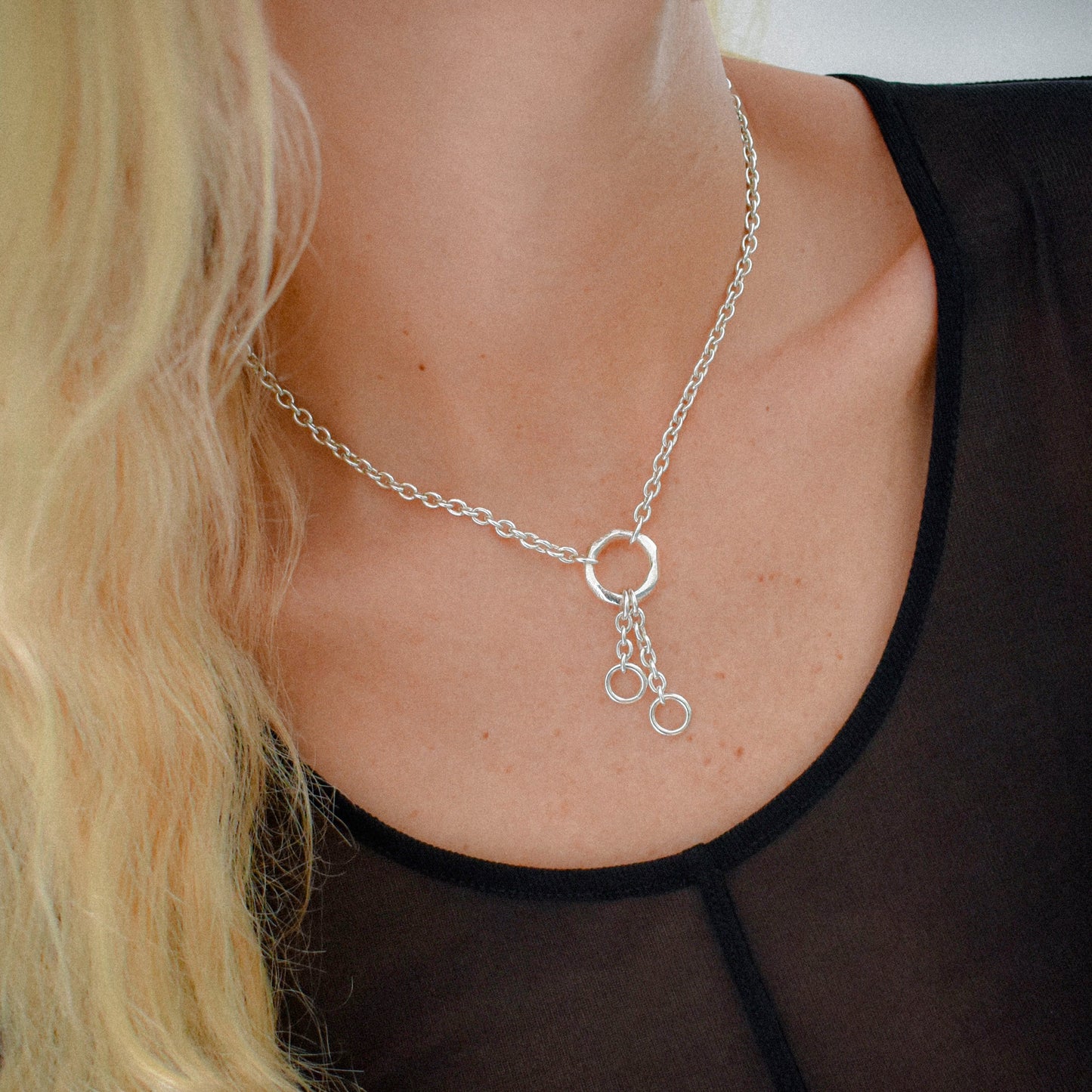 A close-up of a model's neck and chest wearing a silver chain necklace with three interlocking circular ring pendants, styled with a black sheer top.