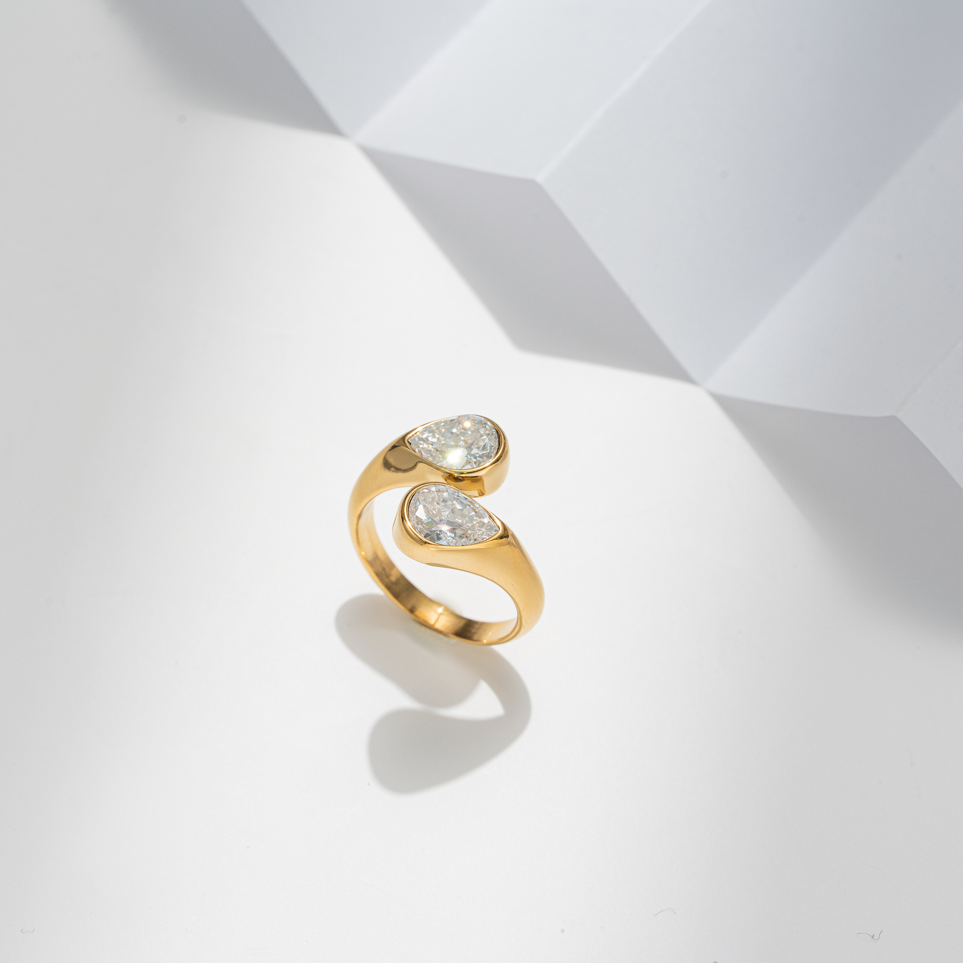 A gold ring with two pear-cut clear gemstones, photographed on a clean white geometric surface with soft shadows.