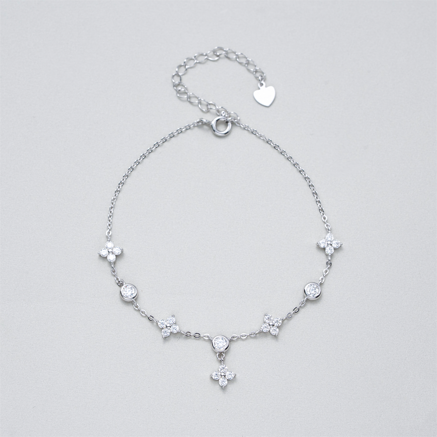 A delicate silver chain anklet featuring five floral starburst charms, three clear round-cut stones, and a small dangling cross charm, displayed on a neutral light gray background.