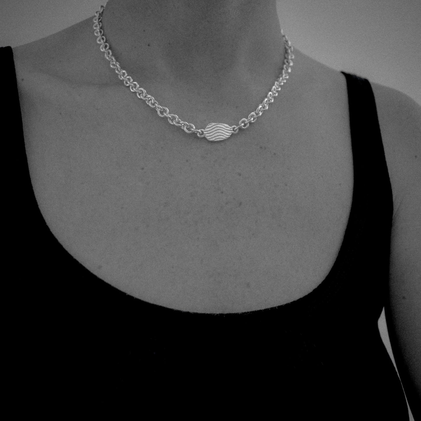 A close-up of a silver chain necklace with a central wave-like charm, worn on a model's neck over a black tank top.