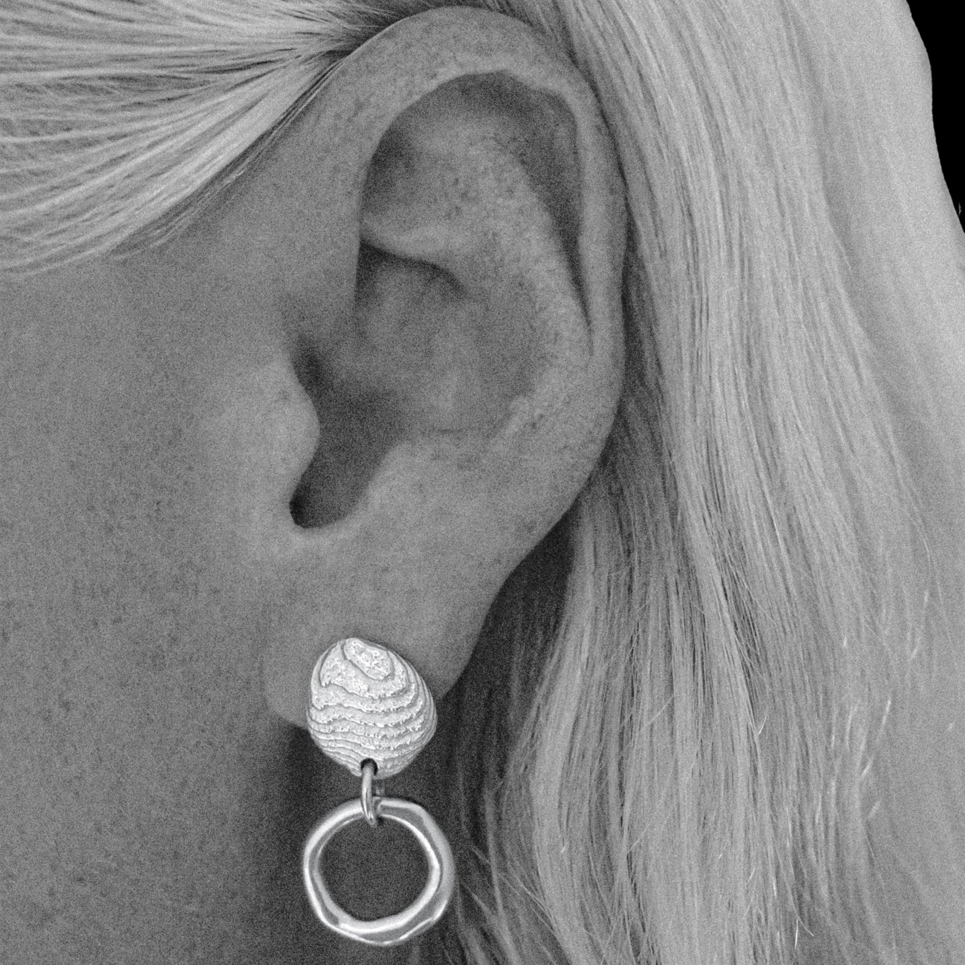 A close-up black and white photograph of a person's ear wearing a textured silver drop earring with a circular dangling element.