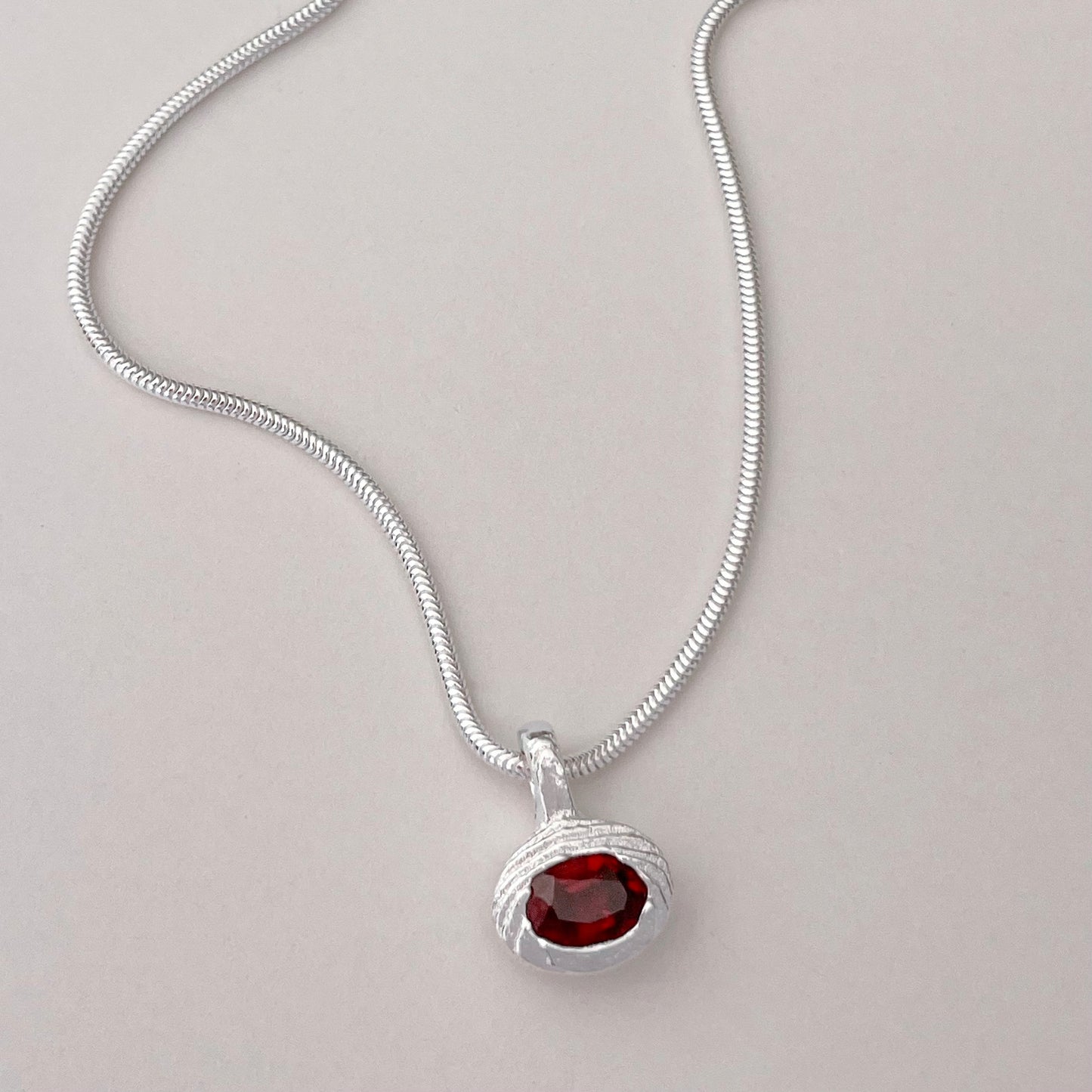 A silver snake chain necklace with a textured silver pendant holding a round red gemstone, displayed on a light gray background.