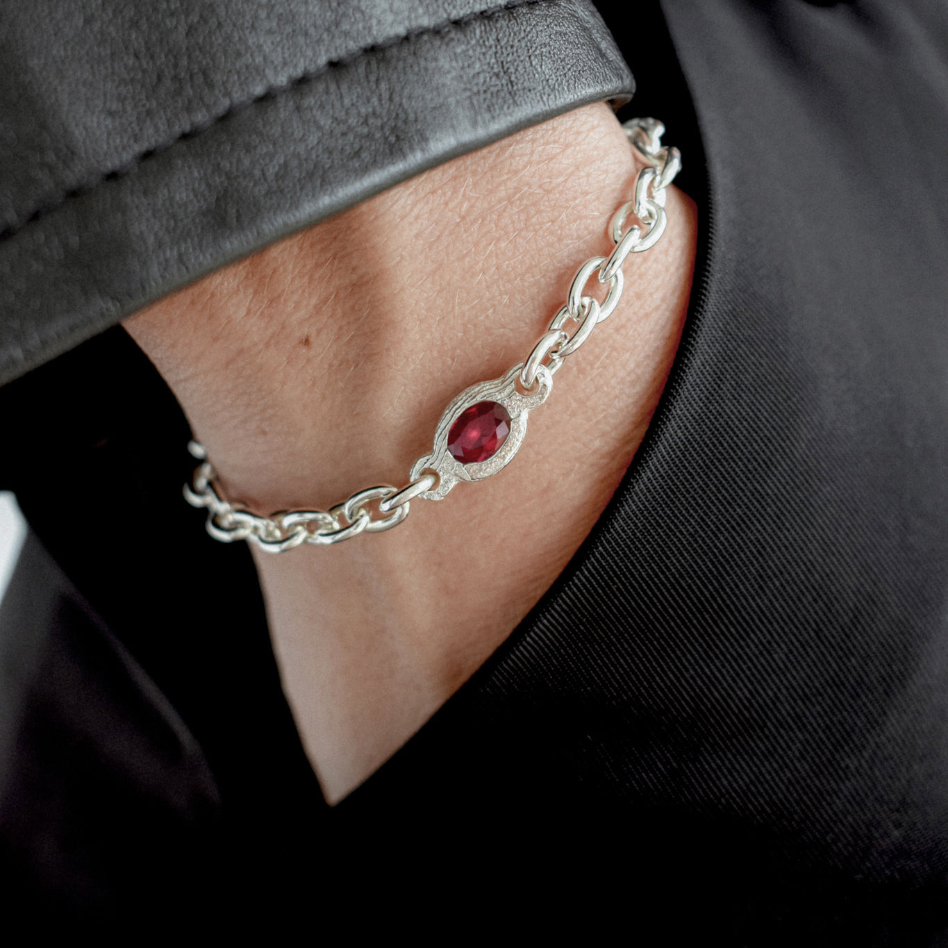 A close-up of a silver chain bracelet with a central oval red gemstone, worn on a person's wrist against a black shirt.