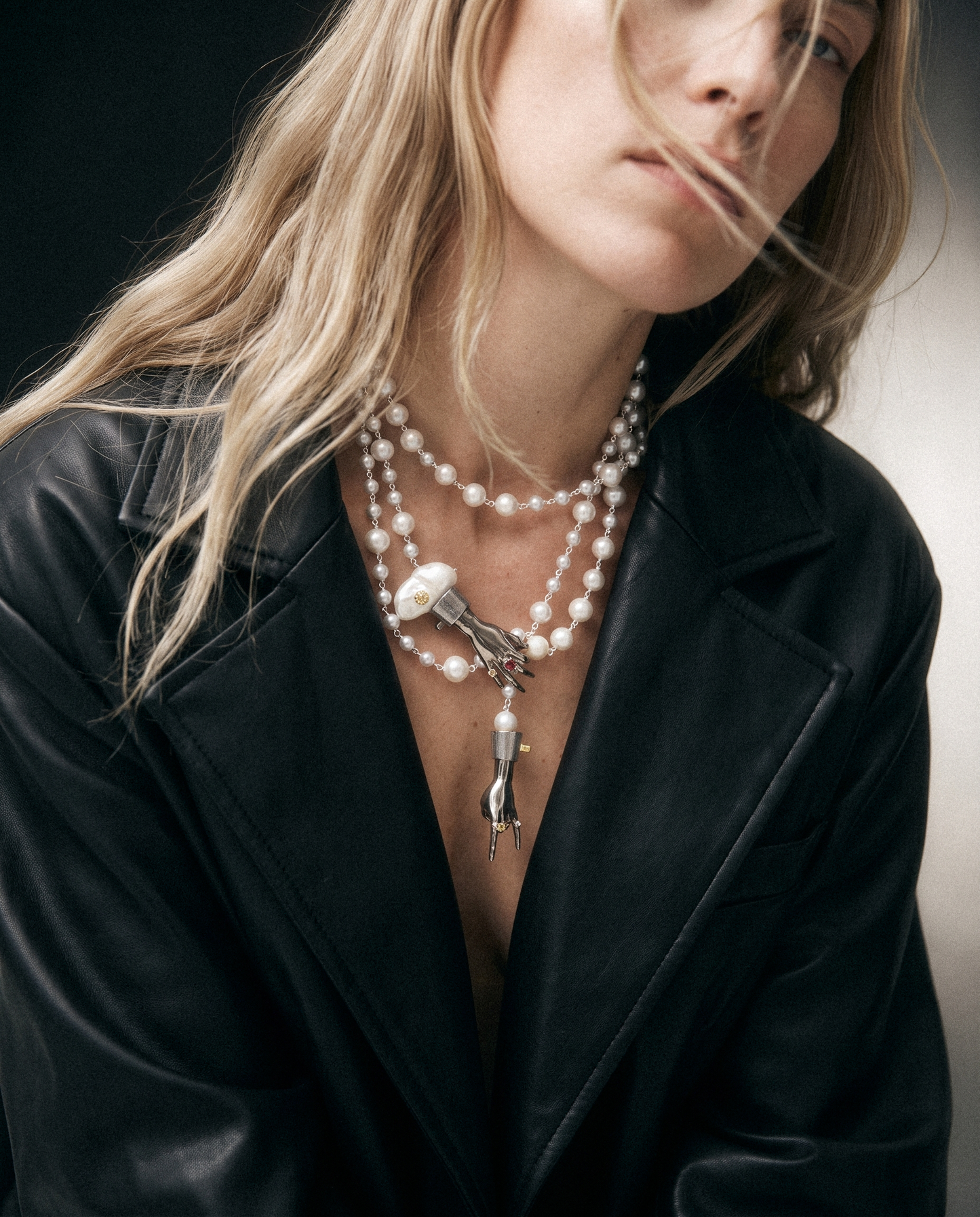A close-up of a woman wearing a black leather jacket and layered pearl necklaces, including one with a silver high-heel charm.