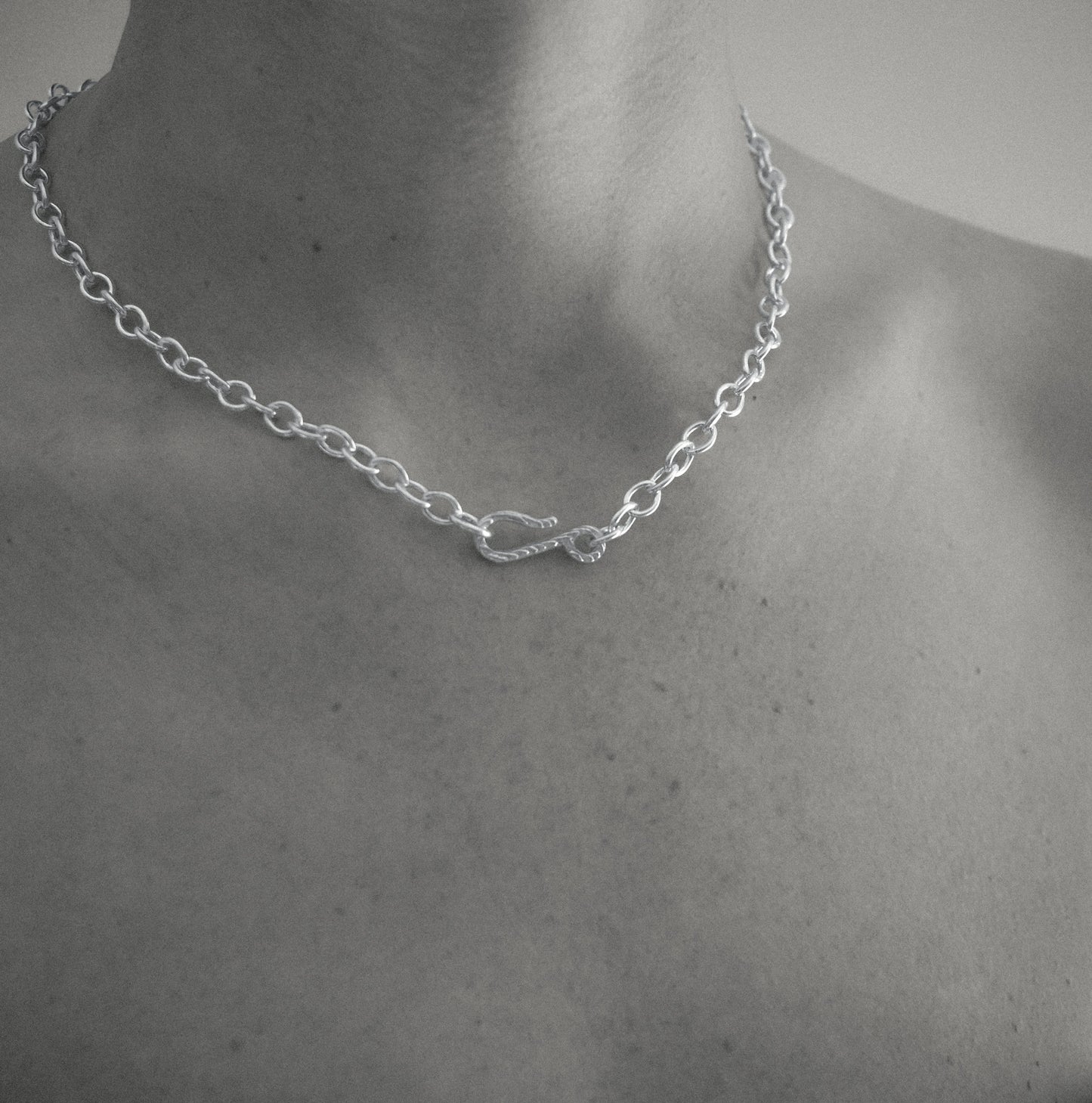 A silver chain necklace with a small infinity-style pendant, shown worn on a person's neck and upper chest in a black-and-white close-up.