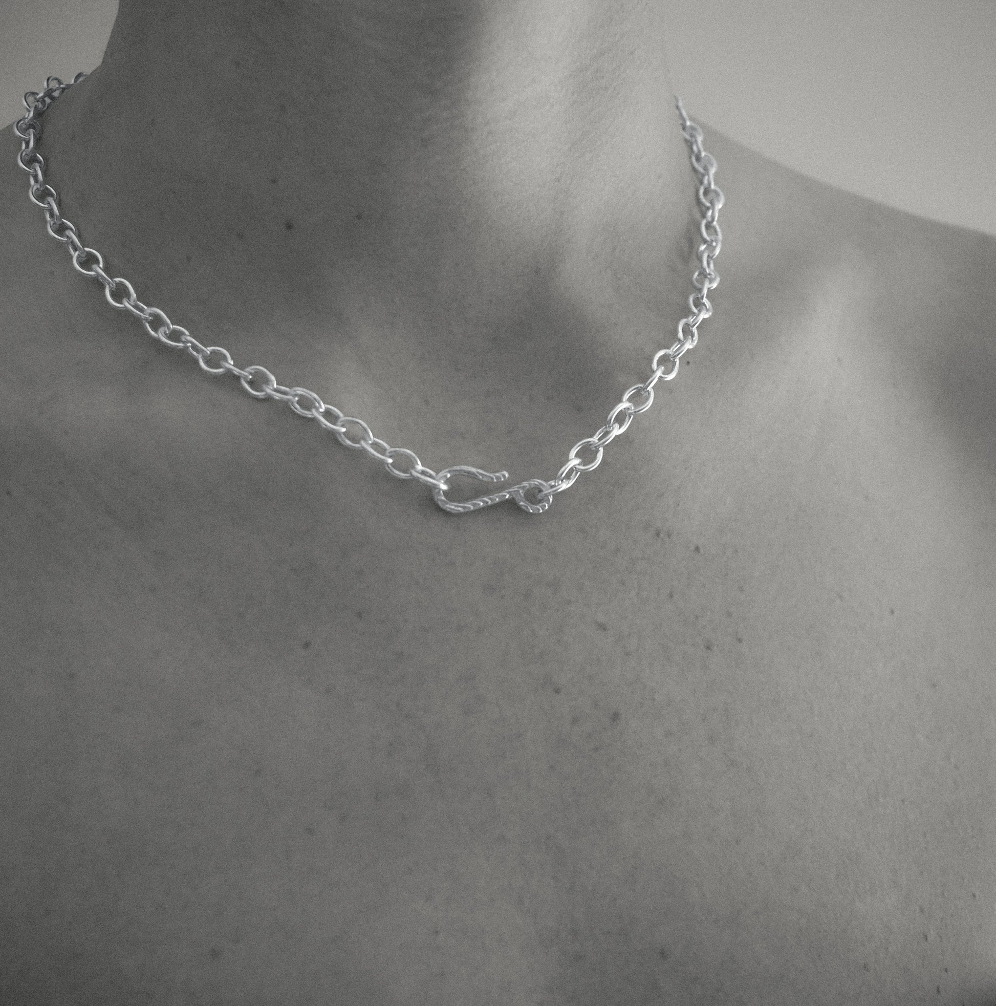 A silver chain necklace with a small infinity-style pendant, shown worn on a person's neck and upper chest in a black-and-white close-up.