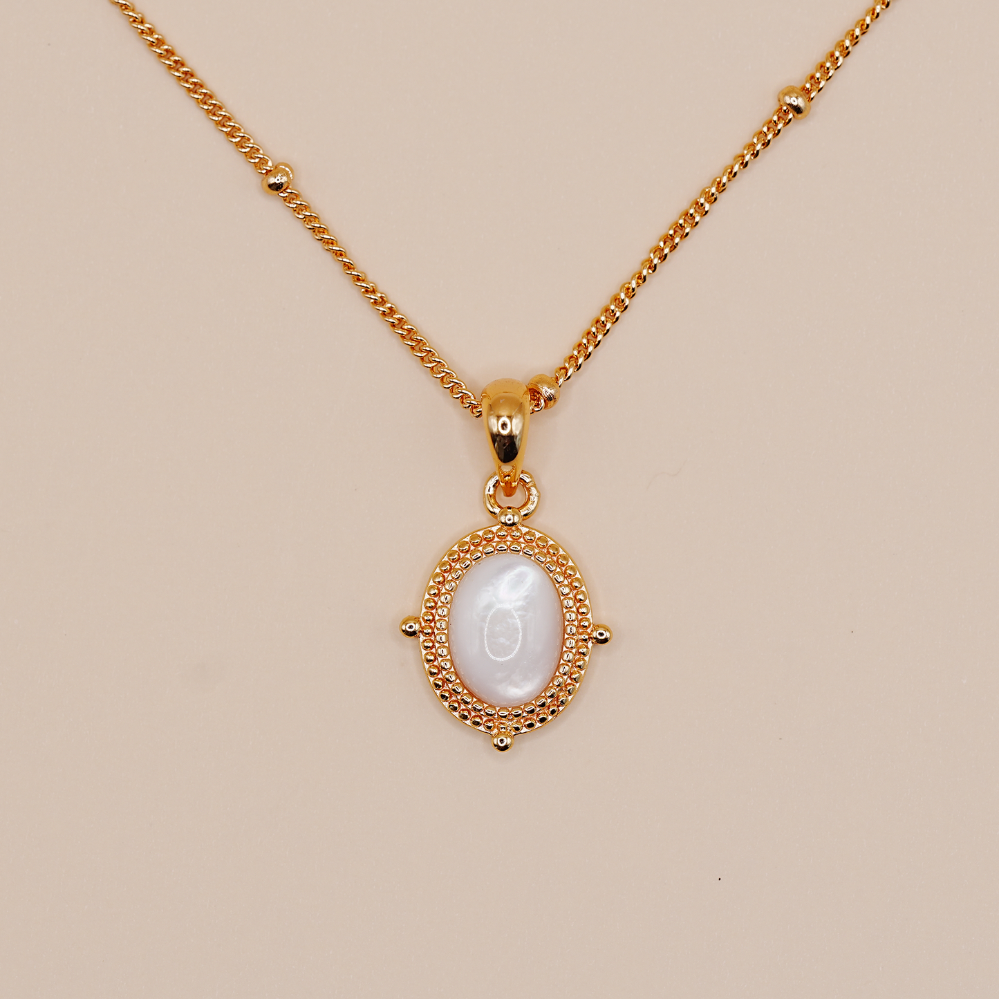 A gold chain necklace with an oval pendant featuring a white mother-of-pearl center and a textured gold border, displayed on a solid beige background.
