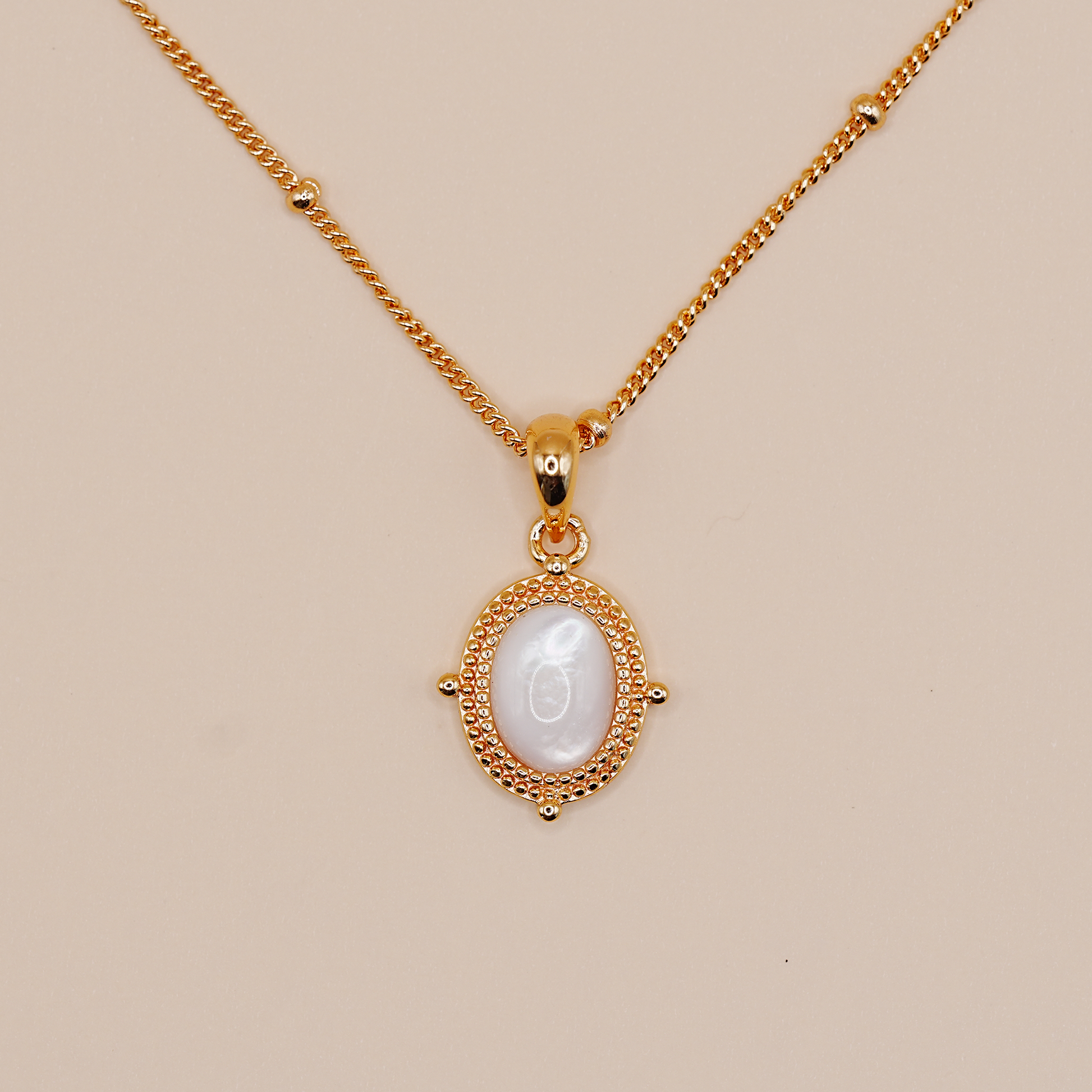 A gold chain necklace with an oval pendant featuring a white mother-of-pearl center and a textured gold border, displayed on a solid beige background.