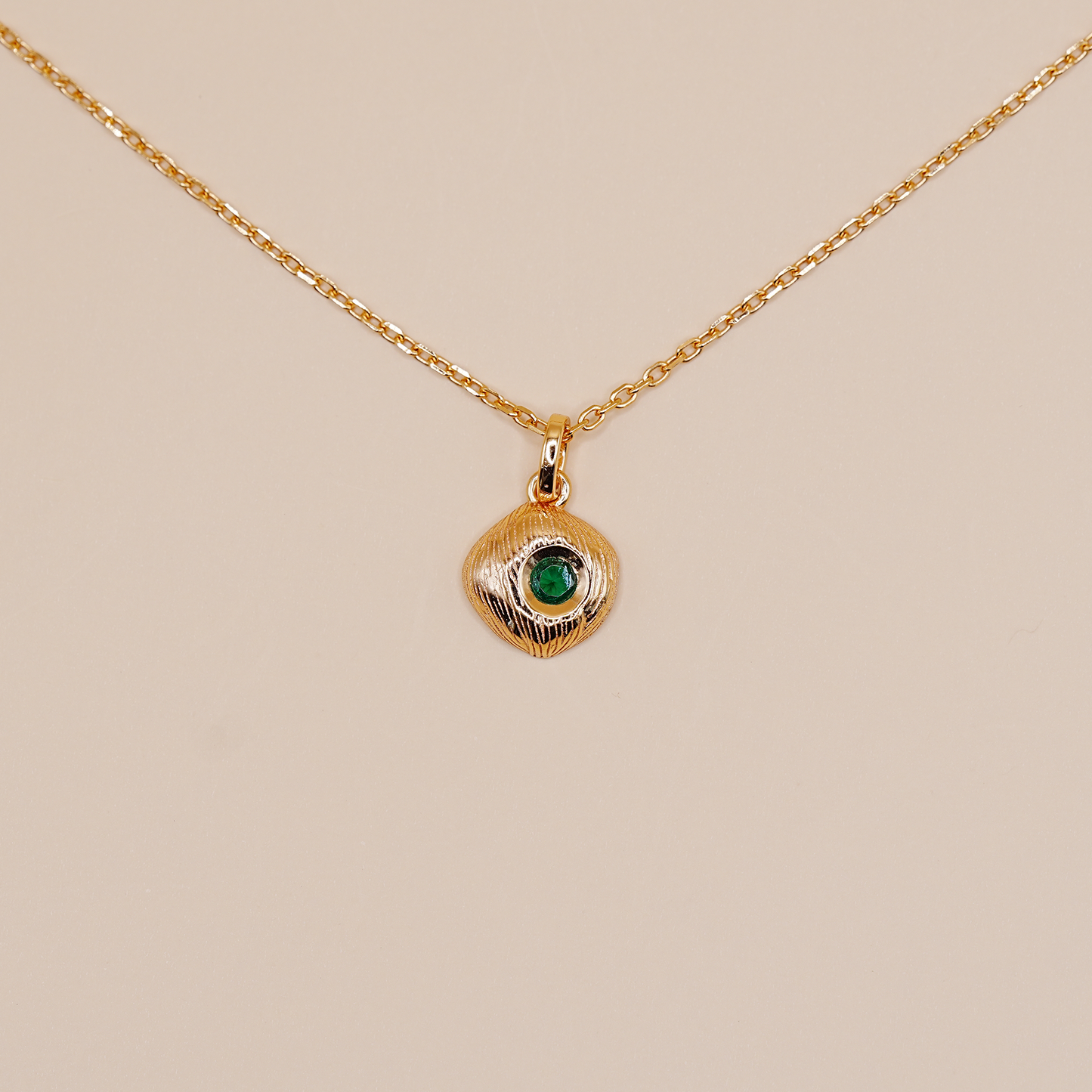 A delicate gold chain necklace featuring a small, textured gold pendant with a central green stone, displayed against a solid beige background.