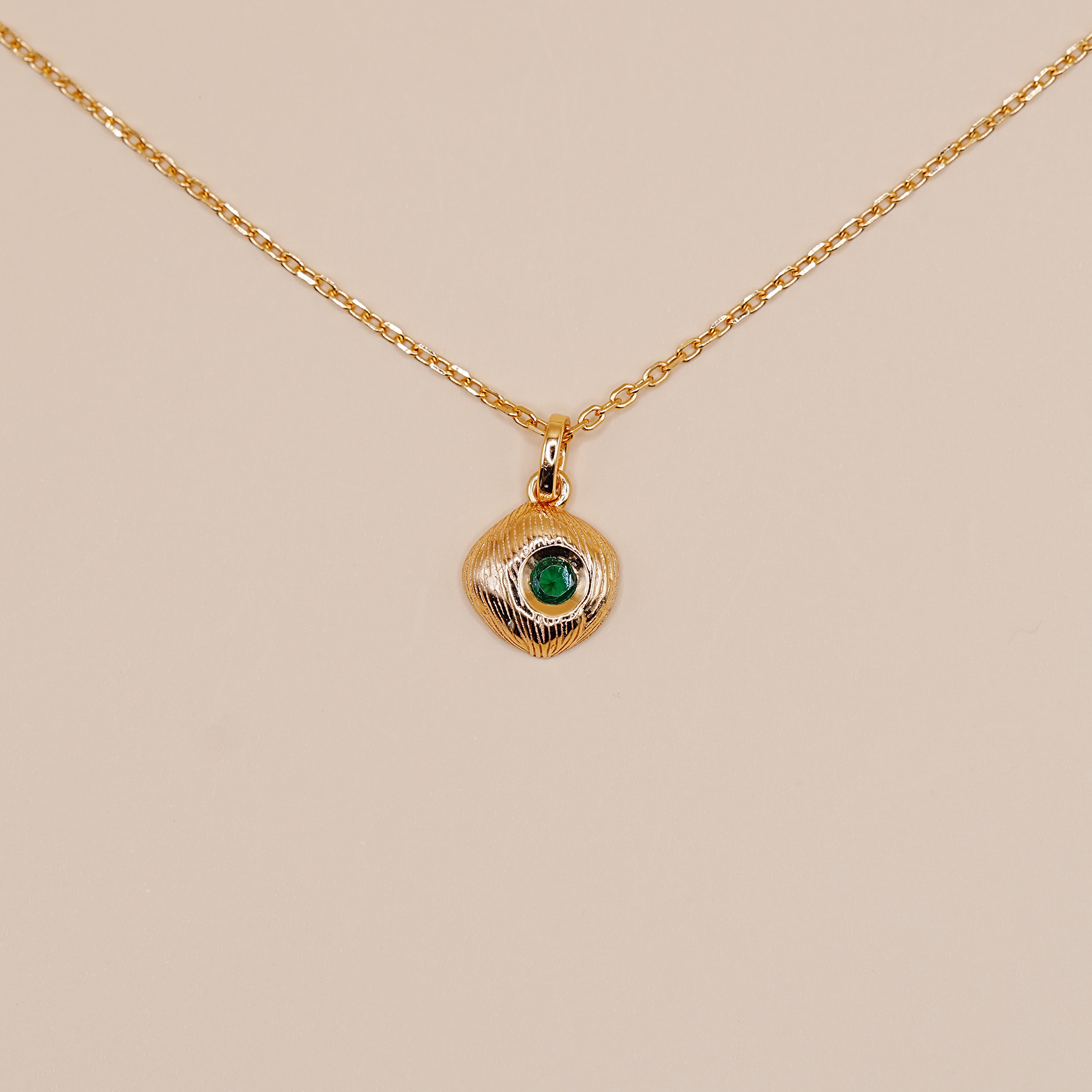 A delicate gold chain necklace featuring a small, textured gold pendant with a central green stone, displayed against a solid beige background.