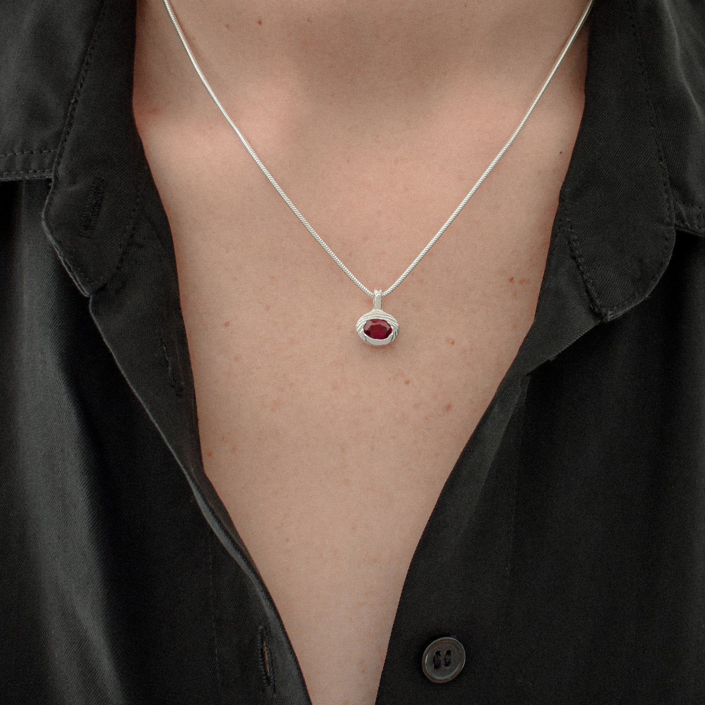 A close-up of a person wearing a delicate silver chain necklace with a small oval red gemstone pendant, styled with an open black button-down shirt.