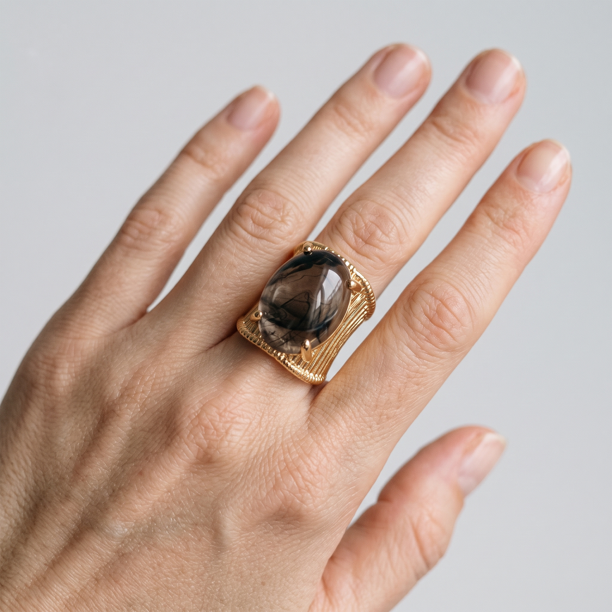 A close-up of a hand wearing a large gold ring with a dark smoky oval gemstone, set against a plain light gray background.