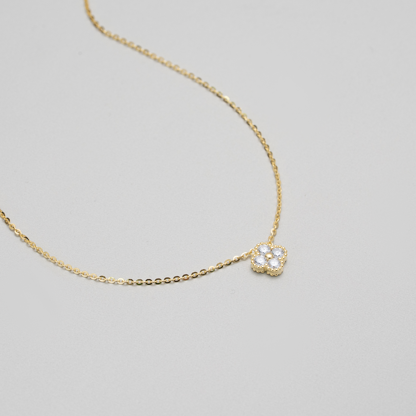 A delicate gold chain necklace featuring a small flower-shaped pendant paved with clear stones, displayed on a neutral light gray background.