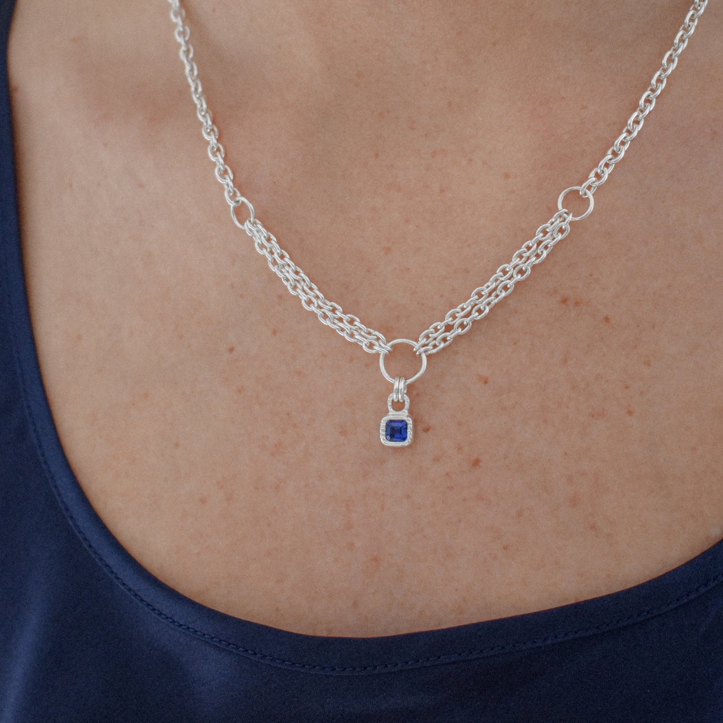 A silver chain necklace with a small square blue gemstone pendant, worn on a model's neck over a navy blue top.