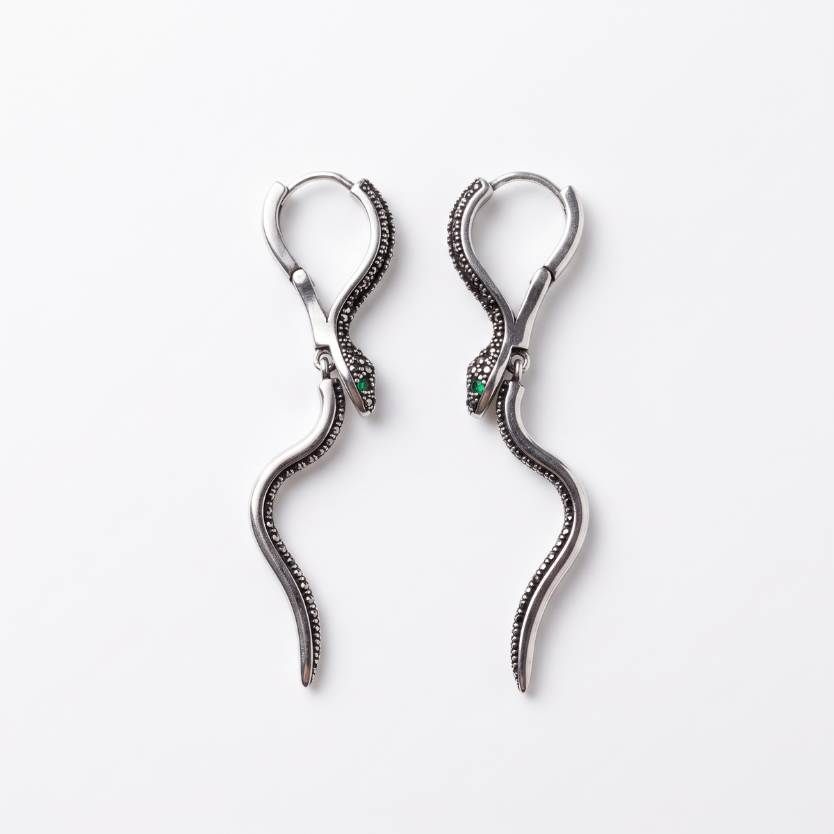 A pair of silver snake drop earrings with textured blackened details and small green gemstone eyes, displayed on a clean white background.