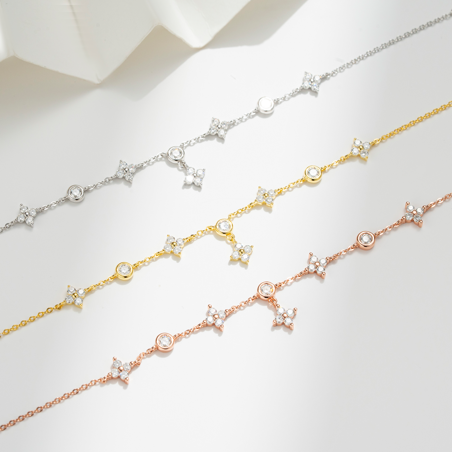 Three delicate chain bracelets in silver, gold, and rose gold, each decorated with small star charms, round bezel-set clear stones, and oval beads, arranged diagonally on a light gray background.