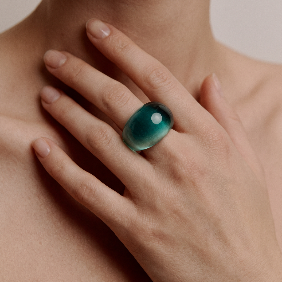 A close-up of a hand wearing a large, smooth, rounded cabochon gemstone ring in a deep teal green color, set against a soft beige background.