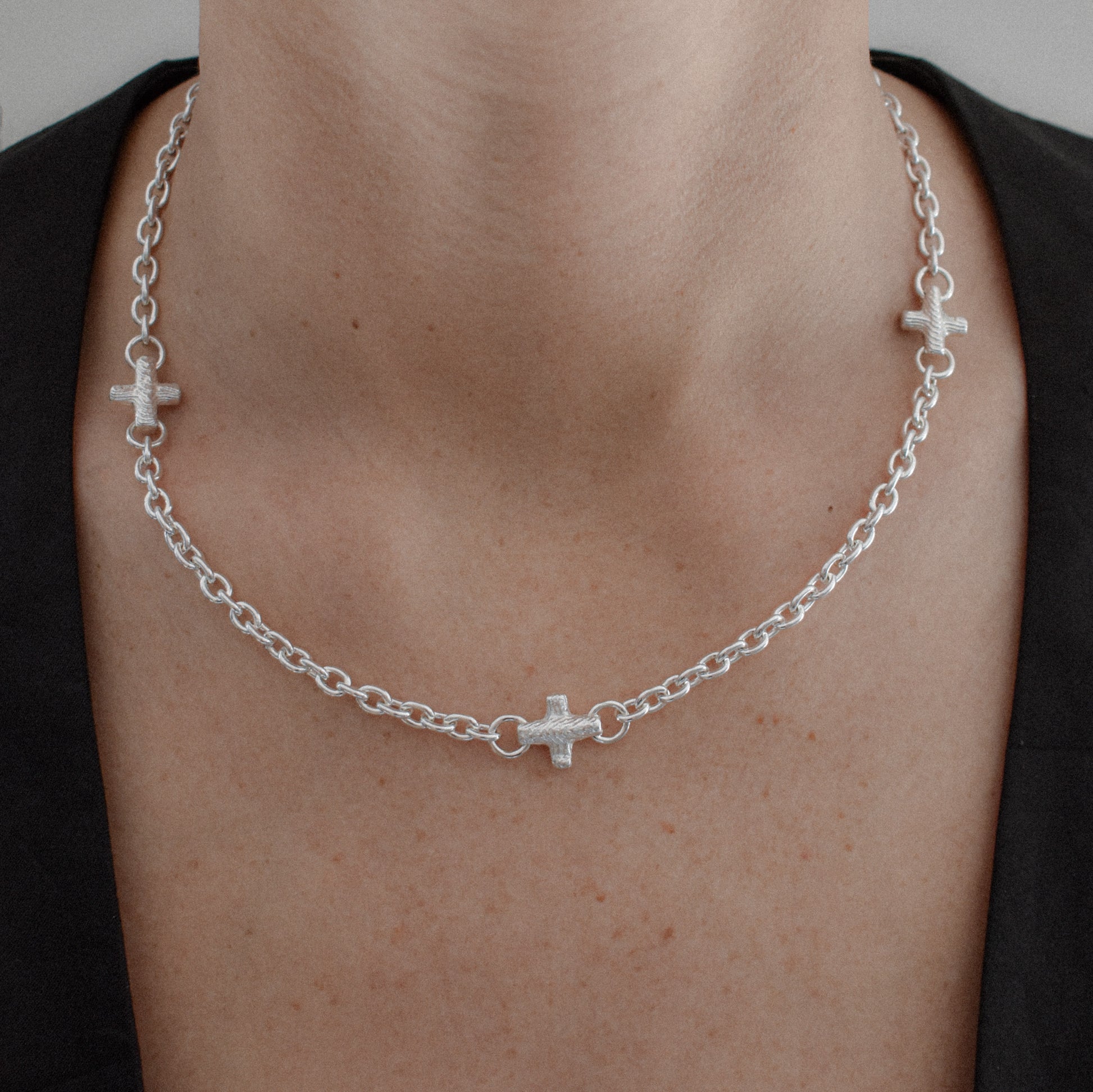 A silver chain necklace with three small cross charms, worn on a person's neck over a black top.