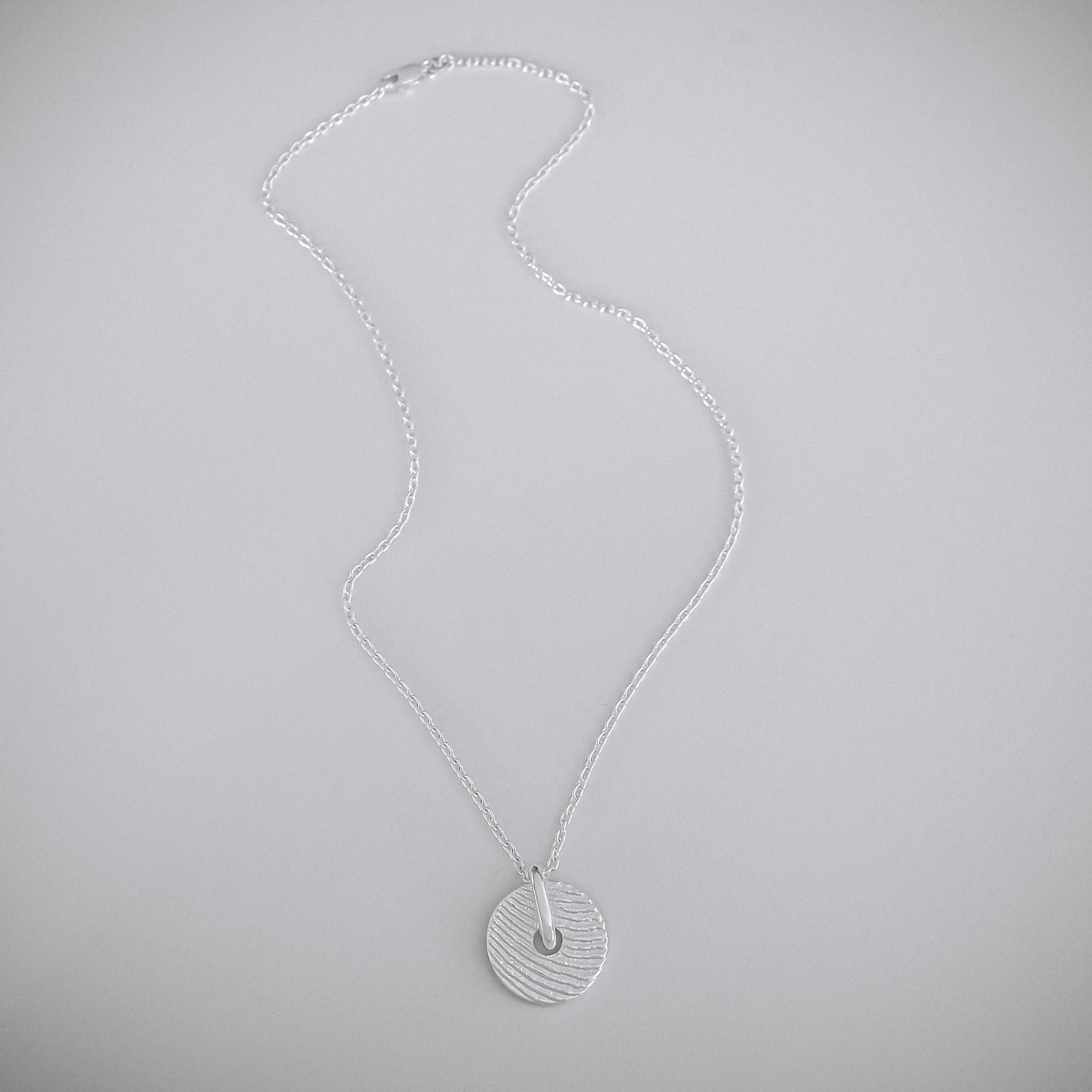 A silver necklace with a textured circular pendant, displayed on a neutral light gray background.