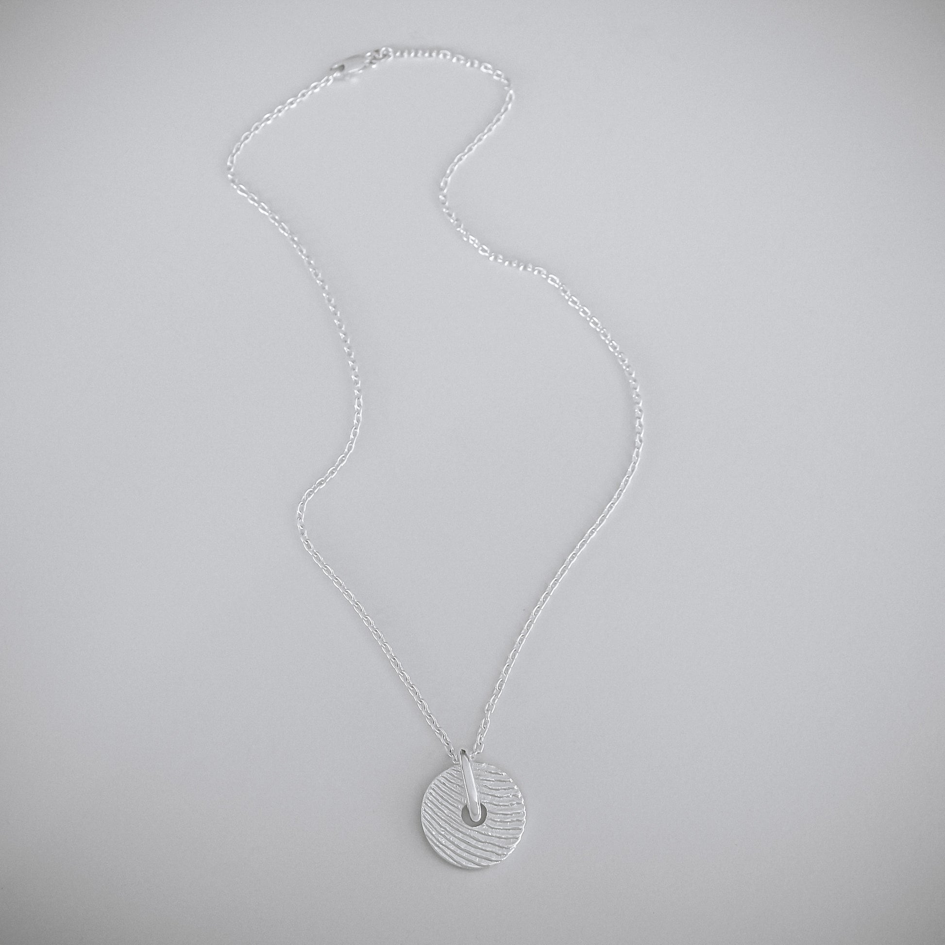 A silver necklace with a textured circular pendant, displayed on a neutral light gray background.