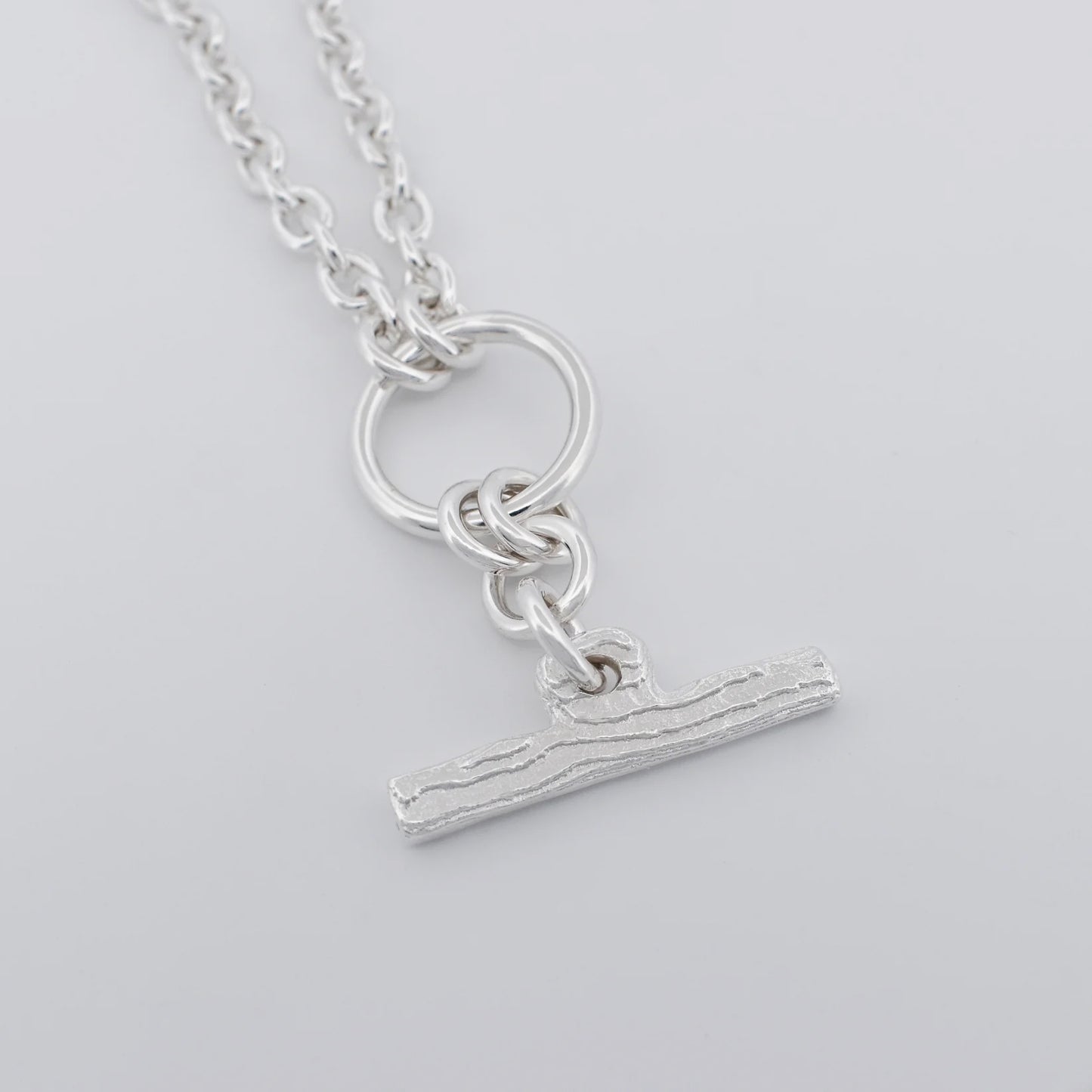 A silver pendant necklace featuring a textured horizontal bar charm suspended from a ring and chain, shown on a light gray background.