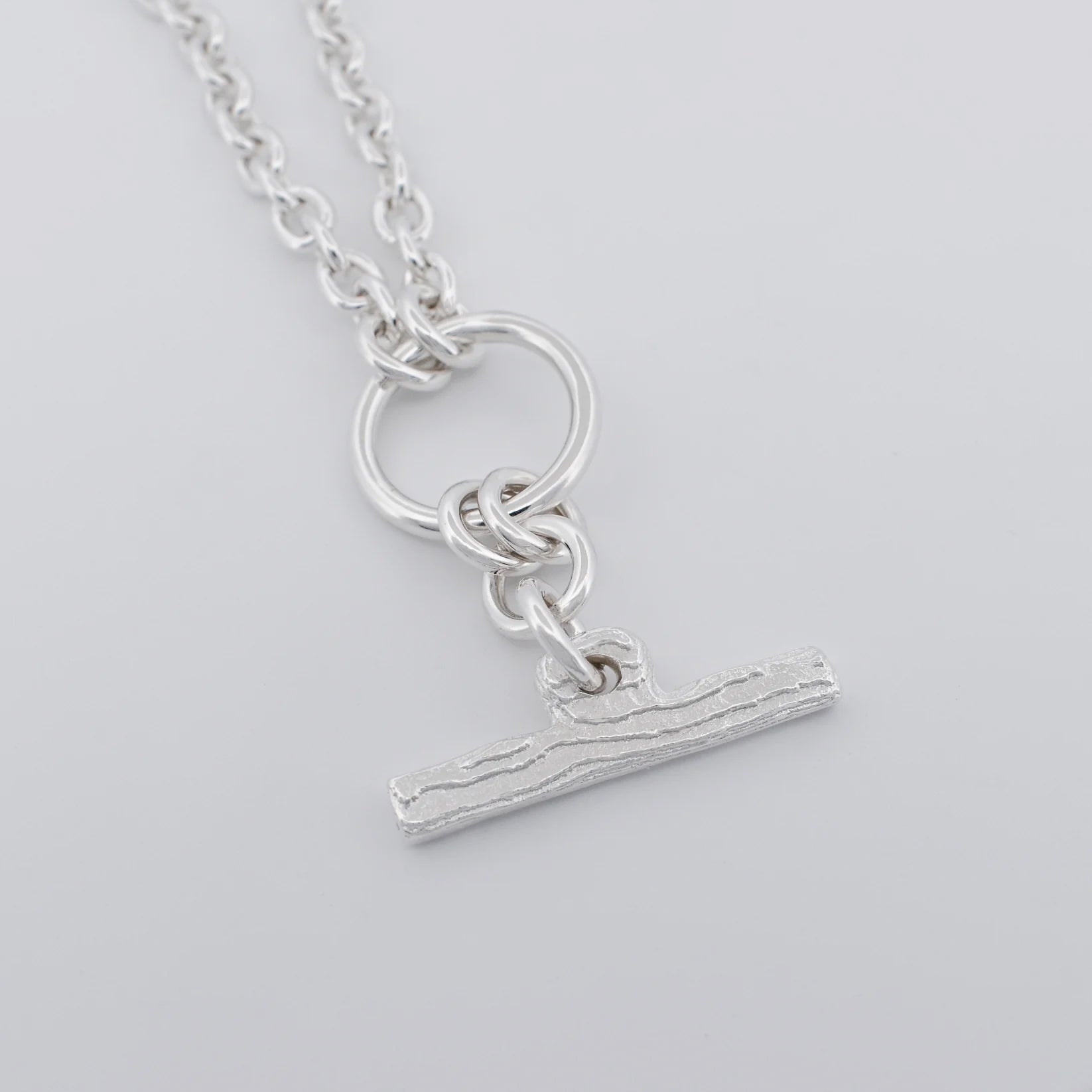 A silver pendant necklace featuring a textured horizontal bar charm suspended from a ring and chain, shown on a light gray background.