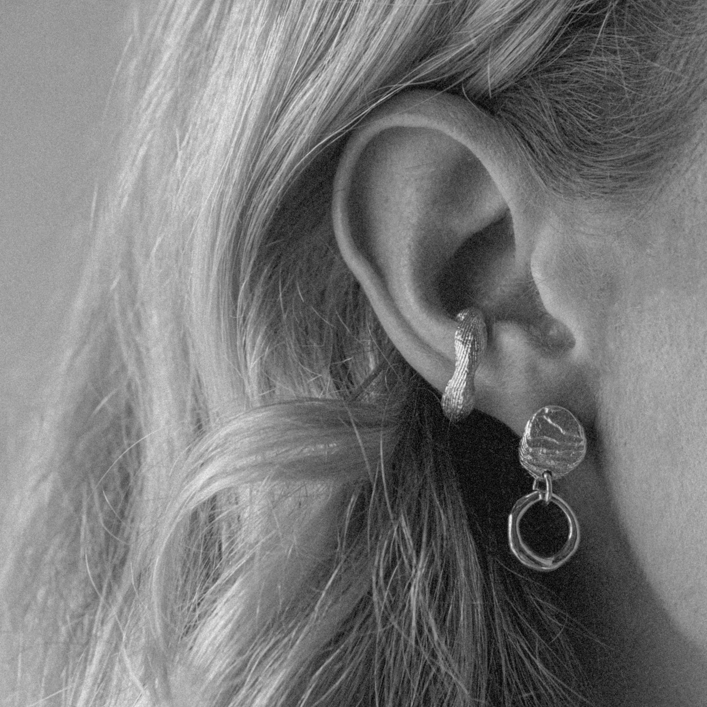 A close-up black and white photograph of a person's ear wearing a textured silver huggie hoop earring with a dangling double-ring earring.
