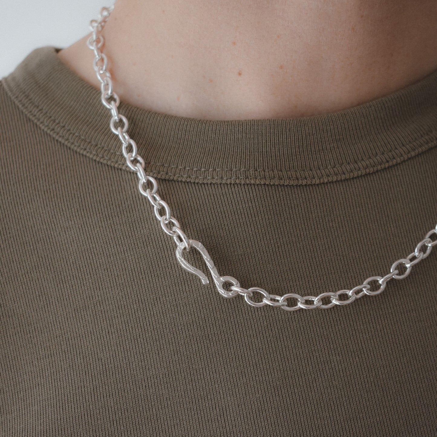 A close-up of a silver chain necklace with a polished infinity symbol pendant, worn on the neck of a person wearing an olive green t-shirt.