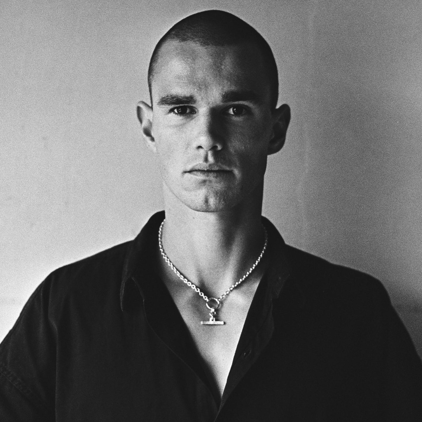 A black and white portrait of a man with a shaved head wearing a dark button-up shirt and a silver necklace with a T-shaped pendant, set against a plain light background.