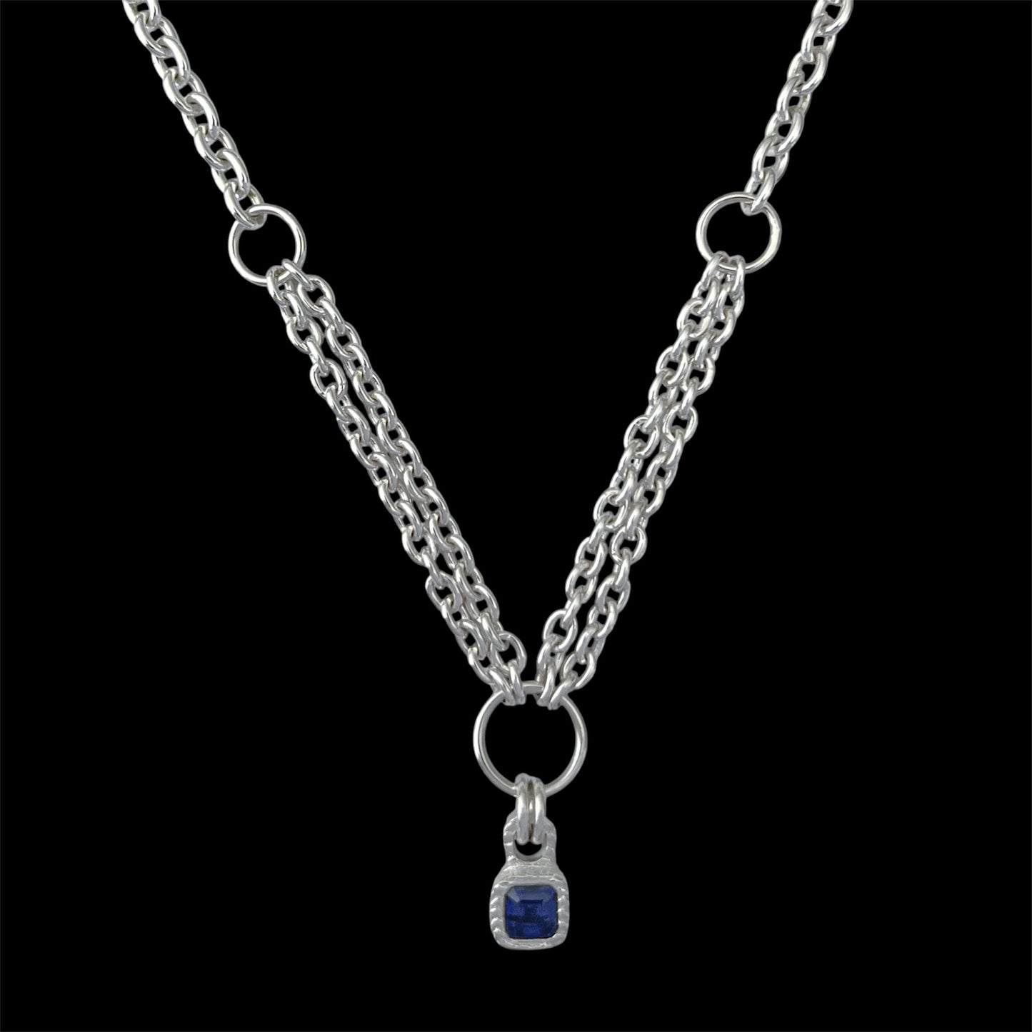 A silver-tone double chain necklace with a small square blue gemstone pendant, displayed against a solid black background.