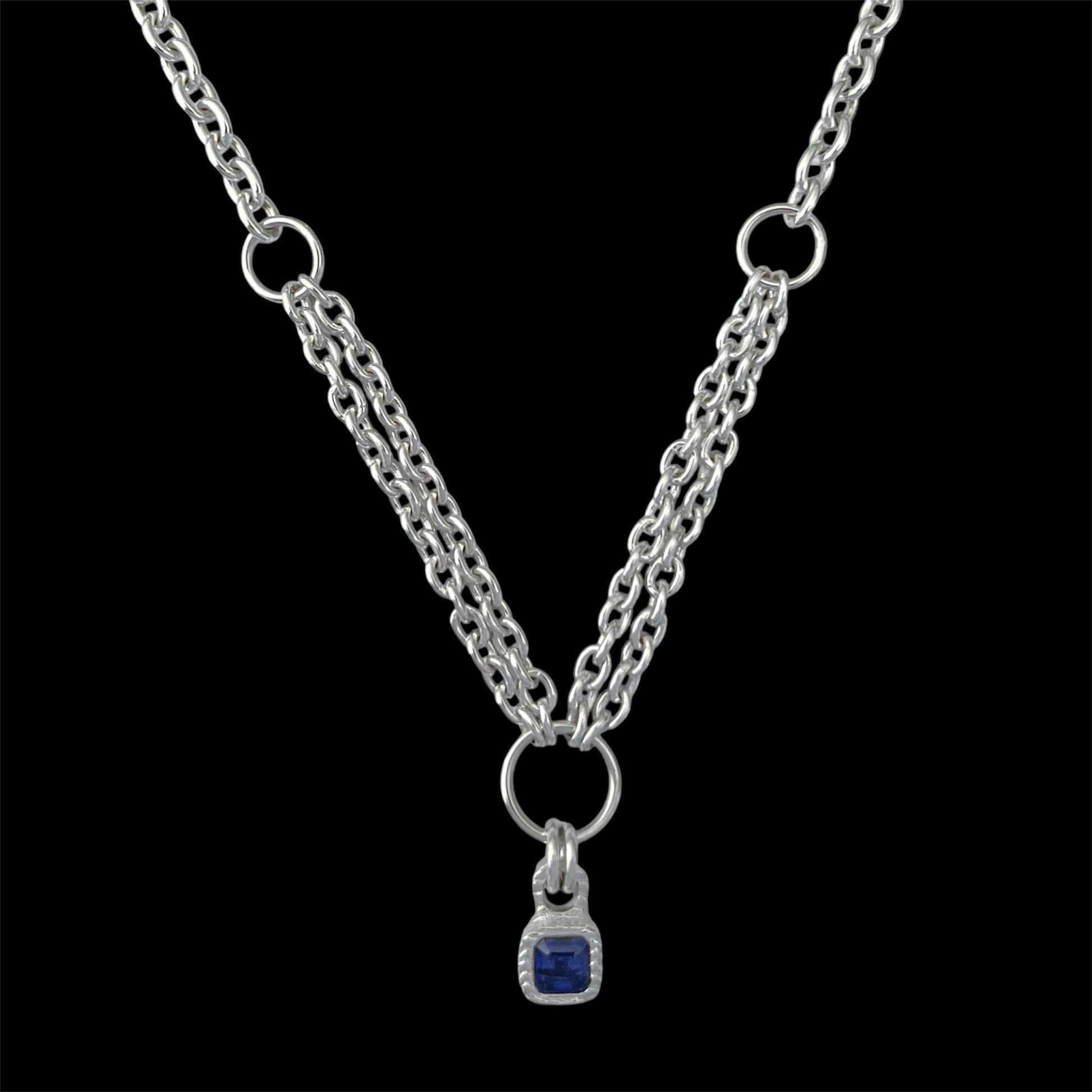 A silver-tone double chain necklace with a small square blue gemstone pendant, displayed against a solid black background.