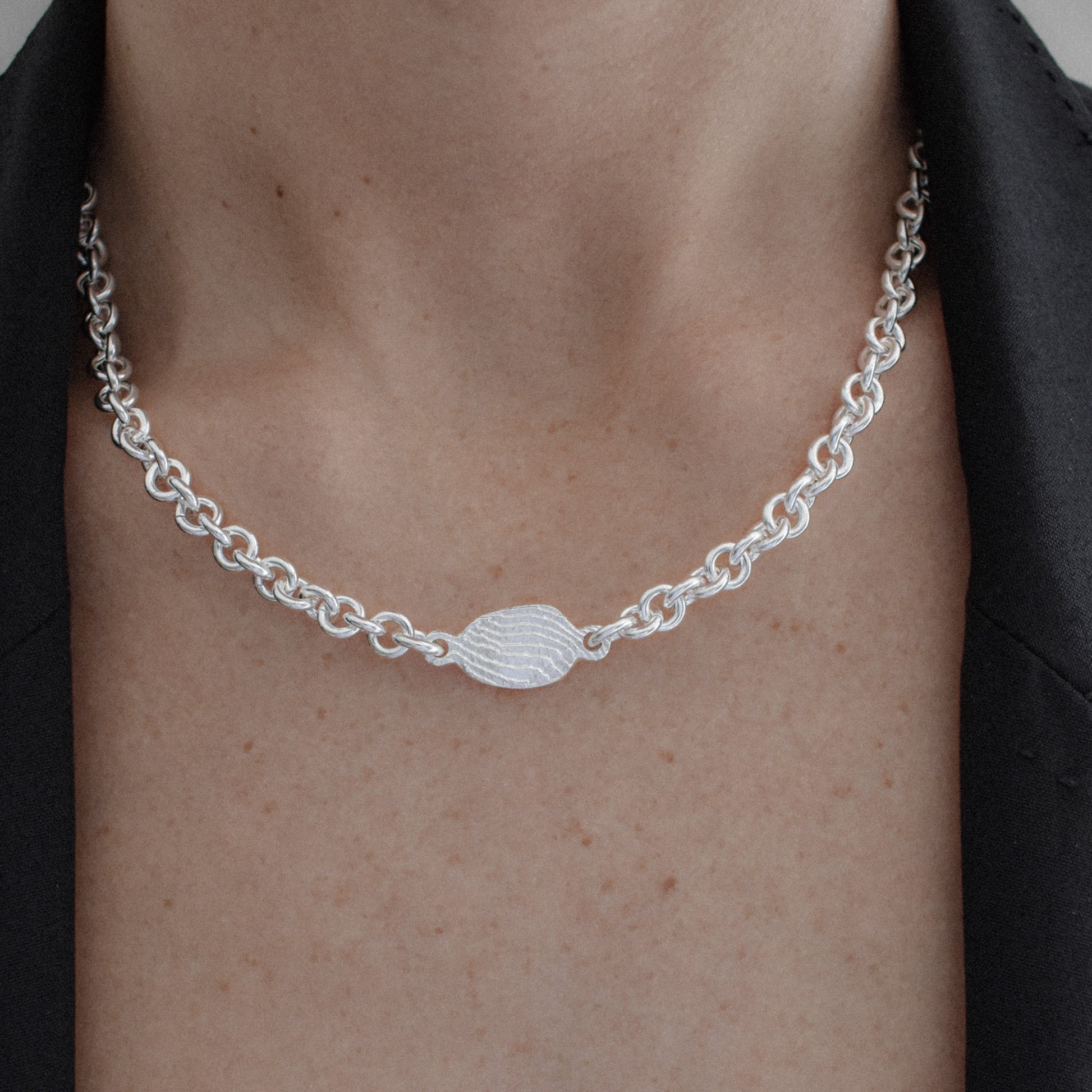 A close-up of a person's neck and chest wearing a silver chain necklace with a textured oval center charm, styled with a black blazer.