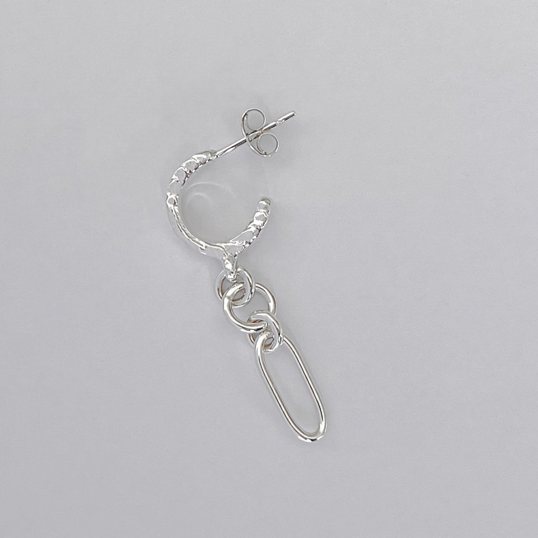 A single silver hoop earring with a textured outer edge, a small drop chain, and a safety-pin style dangling charm, shown on a plain light gray background.
