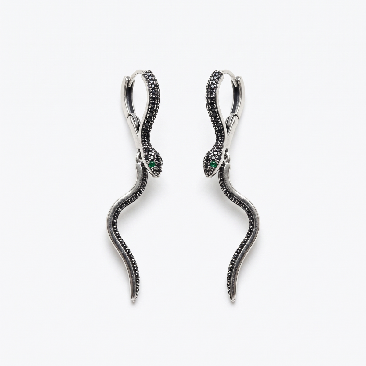 A pair of silver snake drop earrings with black pavé detailing, featuring a small green emerald accent in each eye, set against a plain white background.